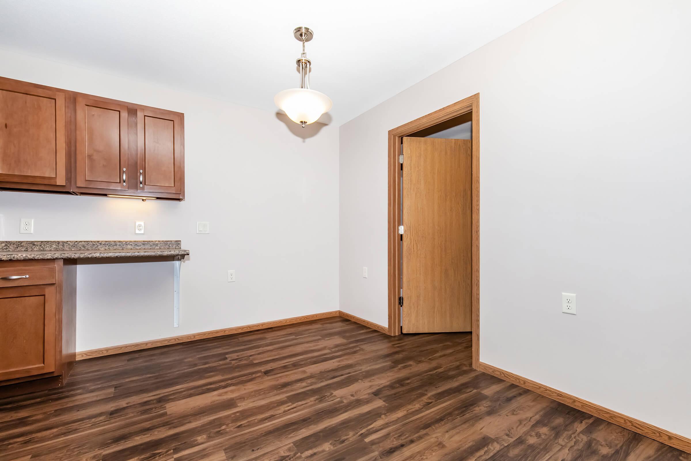 An interior view of a room featuring light brown wooden cabinets, a countertop, and a door that leads to another area. The floor is made of dark wood, and there is a ceiling light fixture. The walls are painted in a neutral color, creating a clean and bright space.