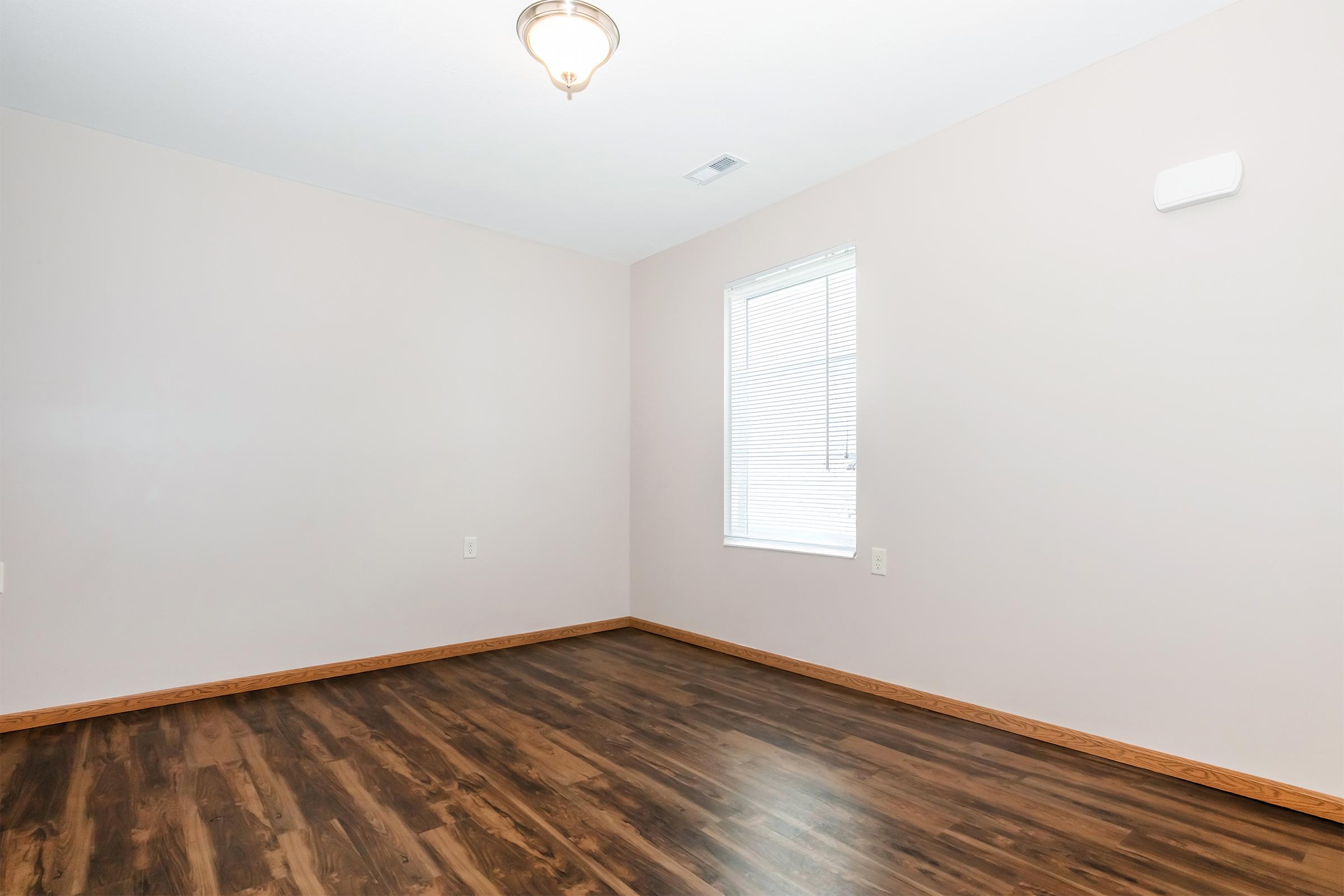 Empty room with light beige walls and dark wooden flooring. A small window with white blinds allows natural light to enter, and a ceiling light fixture hangs in the center. There's a single white wall vent visible on one wall. The space appears simple and uncluttered, ready for decoration or furniture.