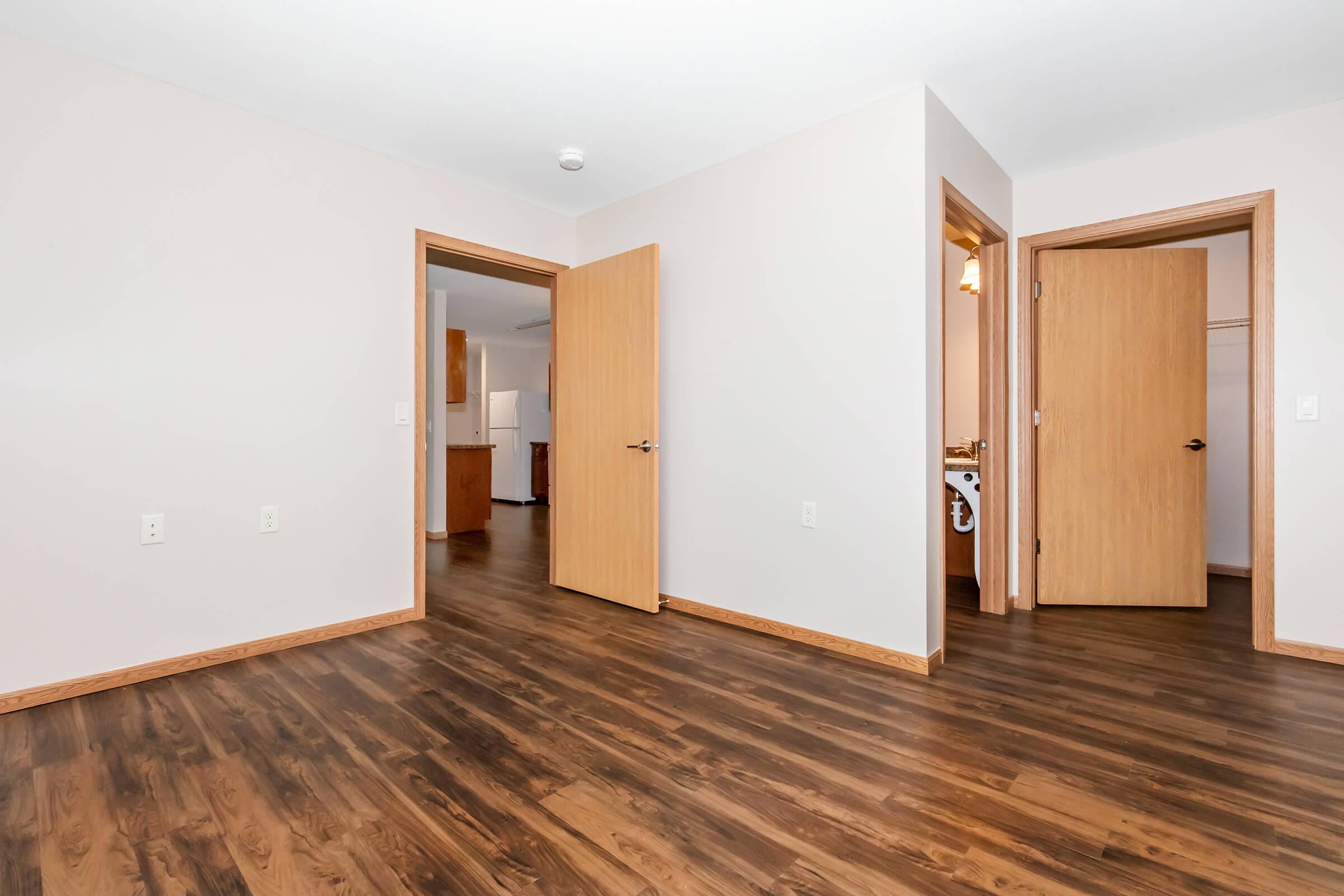 A view of an empty room with light-colored walls and wooden flooring. Two open doors lead to other spaces, suggesting a layout with multiple rooms. Natural light may enter from unseen windows, creating a bright atmosphere. The overall setting appears modern and welcoming, ideal for various uses.