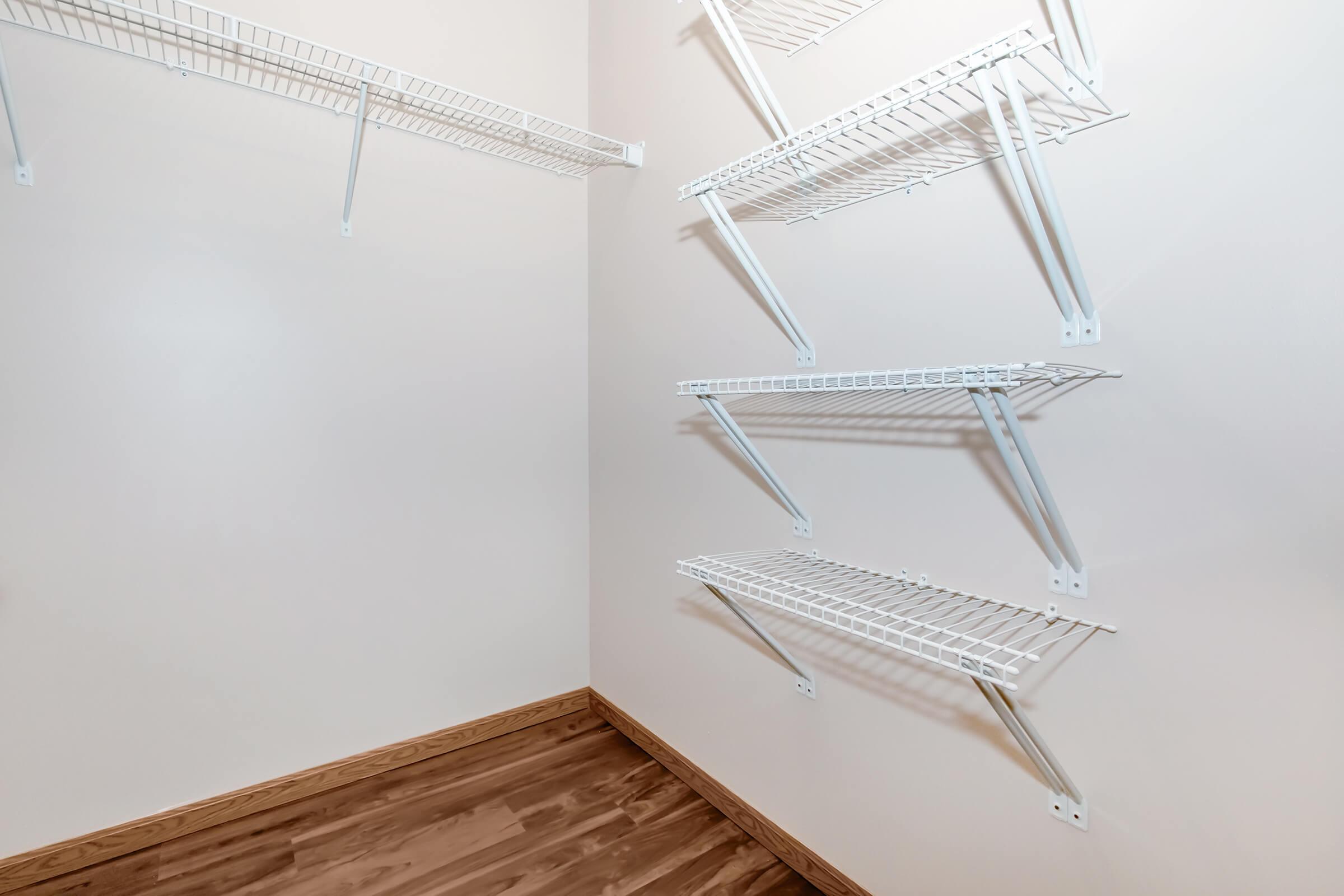 A clean, empty closet featuring white wire shelving arranged on two walls. The floor is made of light-colored wood, creating a warm contrast with the shelving and walls. The closet has a bright, neutral-colored finish, providing a spacious and organized appearance.