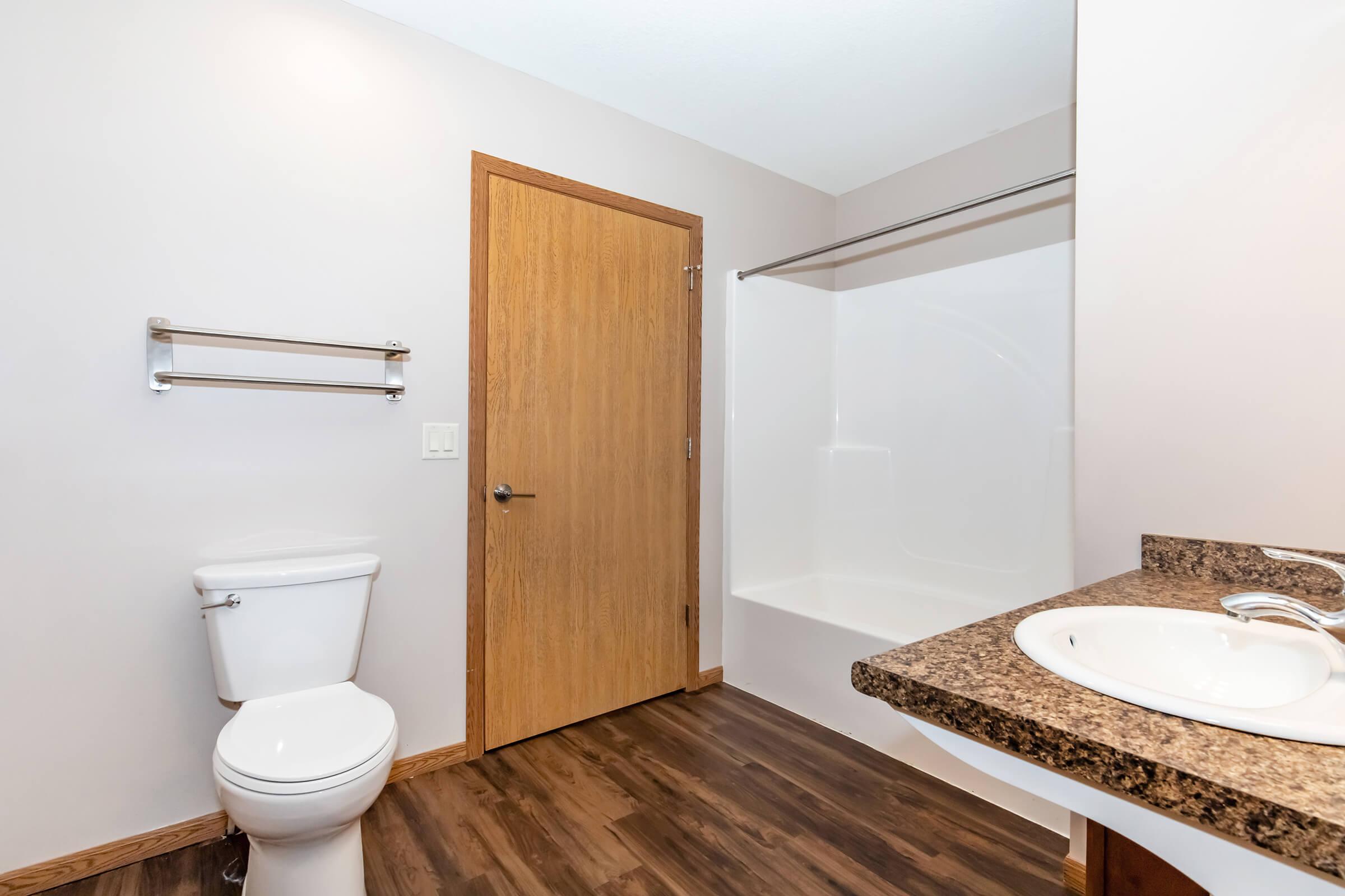 A clean, modern bathroom featuring a white toilet, a bath/shower area with a smooth wall, a granite countertop with a sink, a towel rack on the wall, and a wooden door leading out. The flooring is wooden, enhancing the overall neutral and contemporary aesthetic.