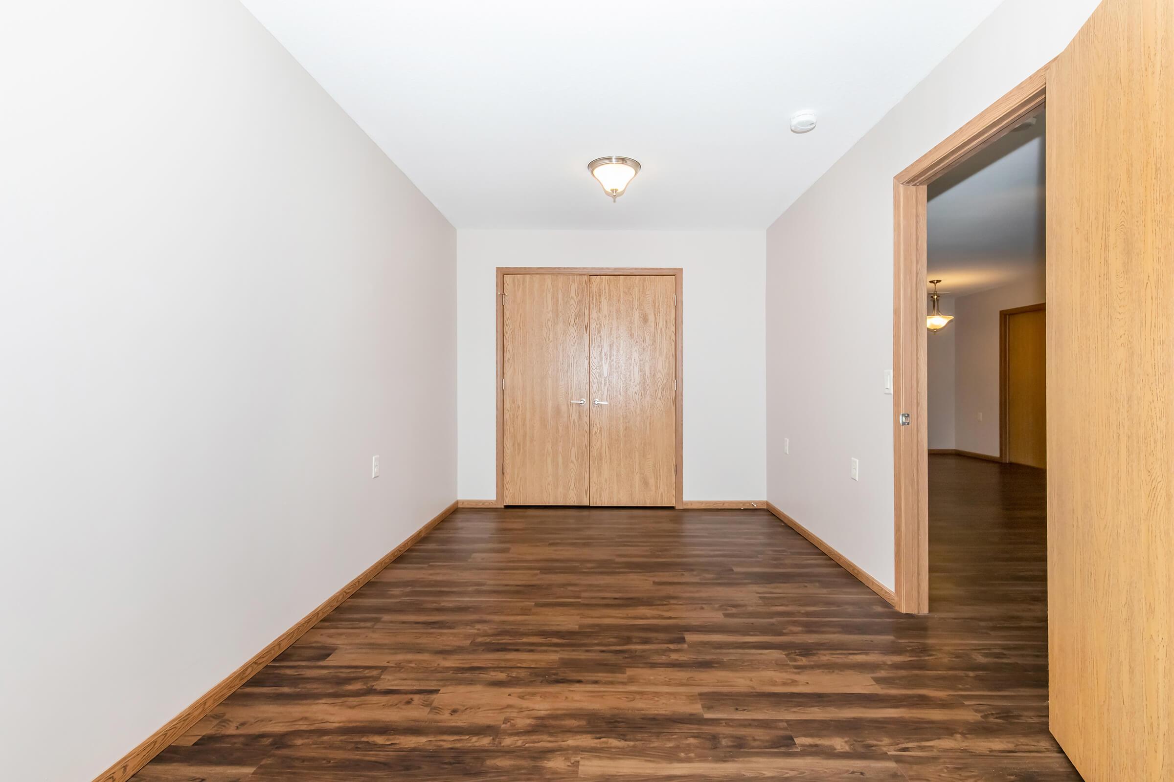 A spacious hallway with light-colored walls and dark wooden flooring. There is a closed wooden door on the far wall and an open doorway to the right leading to another room. Ceiling lights illuminate the area, creating a warm and inviting atmosphere.