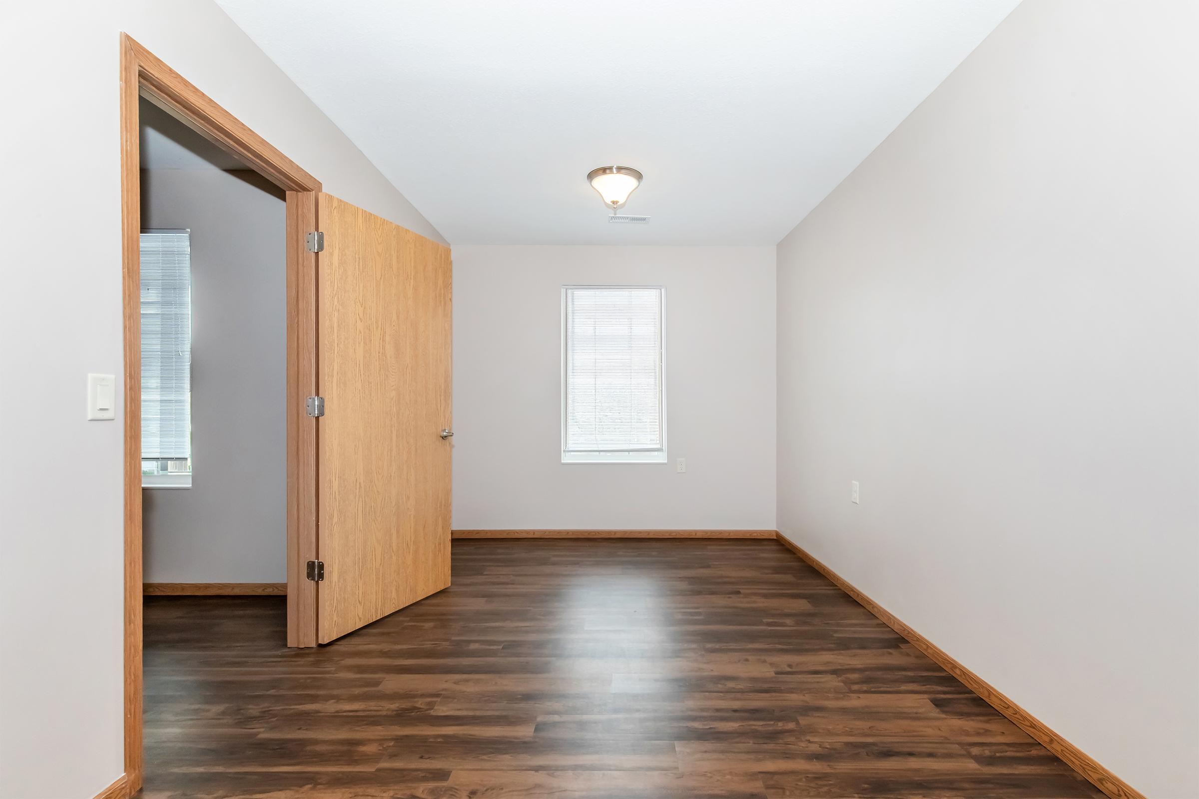 Interior view of a room featuring light gray walls and medium wood flooring. A partially open wooden door leads to another space, and there is a window with blinds. The ceiling is fitted with a simple light fixture, creating a bright and airy atmosphere.