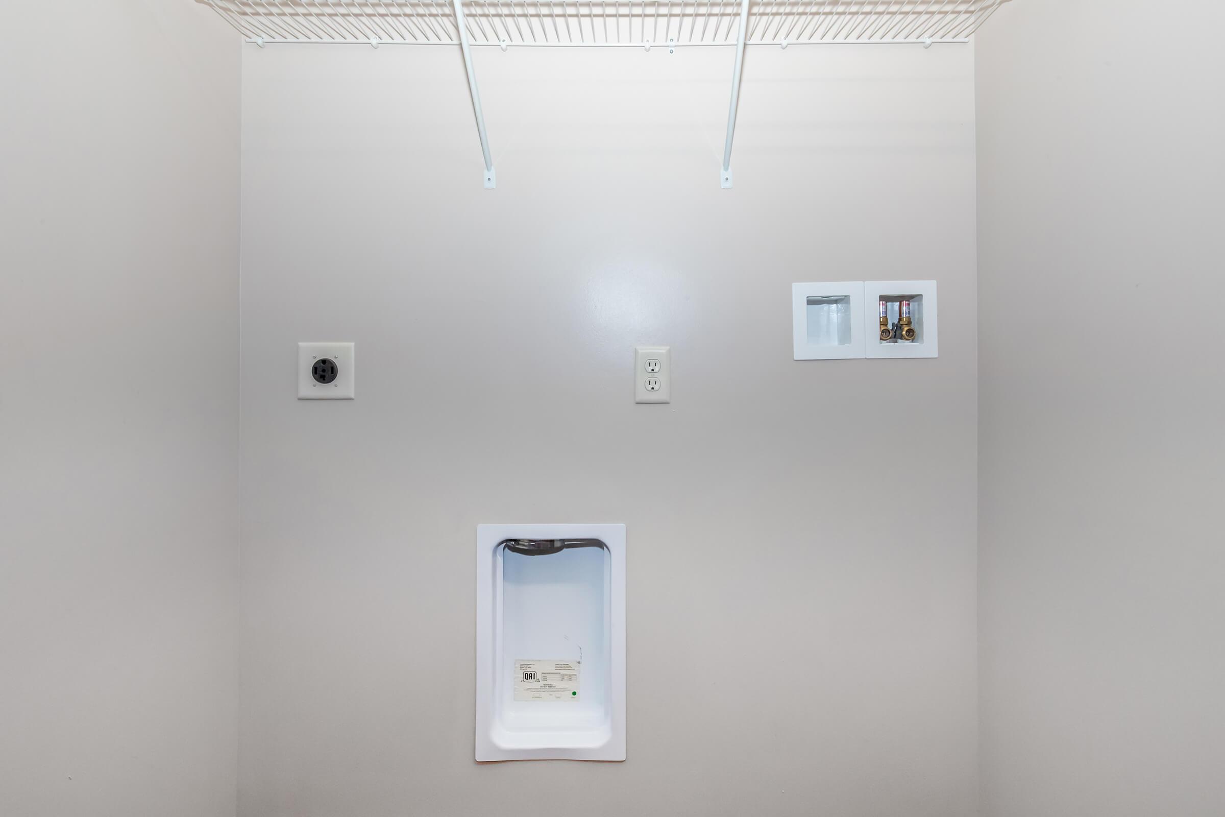 A blank laundry room space featuring a white wall with a hanging shelf. There is an electrical outlet on the left side, a circuit breaker panel on the right, and a small white unit installed in the wall at the center, likely for a washing machine or water connection. The area is well-lit and uncluttered.