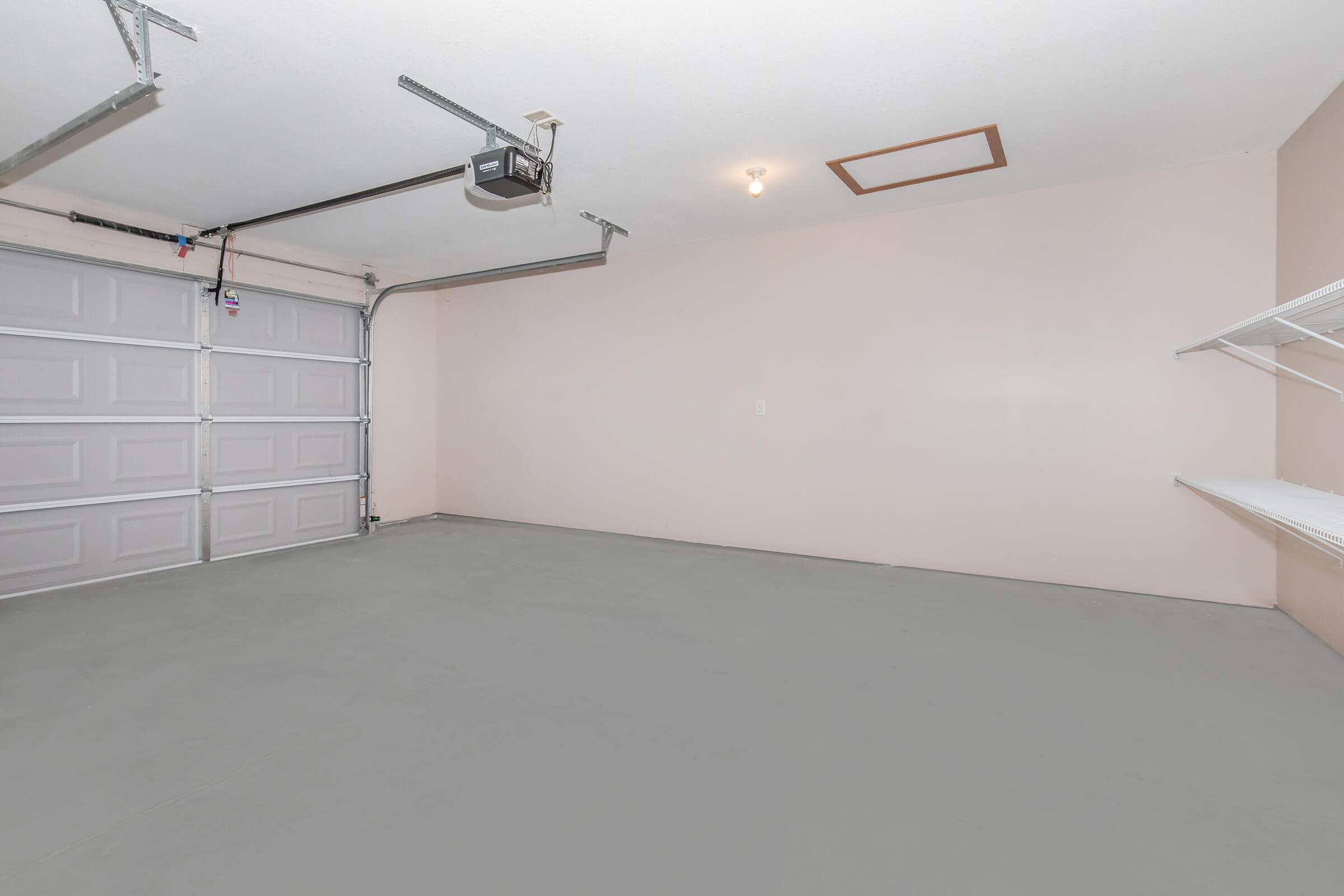 A clean, empty garage with a light gray floor and pink walls. The garage door is closed, and a ceiling light fixture is visible. A wall-mounted shelf is installed on the right side, providing storage space. The overall space appears well-maintained and organized, ready for use.