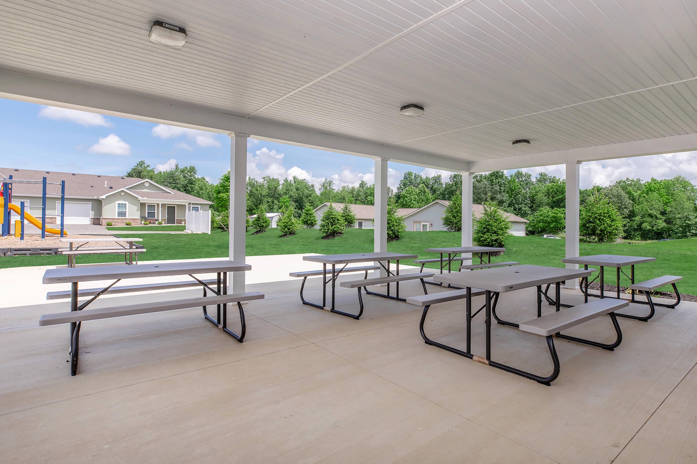Covered outdoor seating area with multiple picnic tables, surrounded by green grass and trees, and a playground visible in the background. The space is bright and spacious, under a white ceiling with overhead lights. A few houses are also seen beyond the seating area.