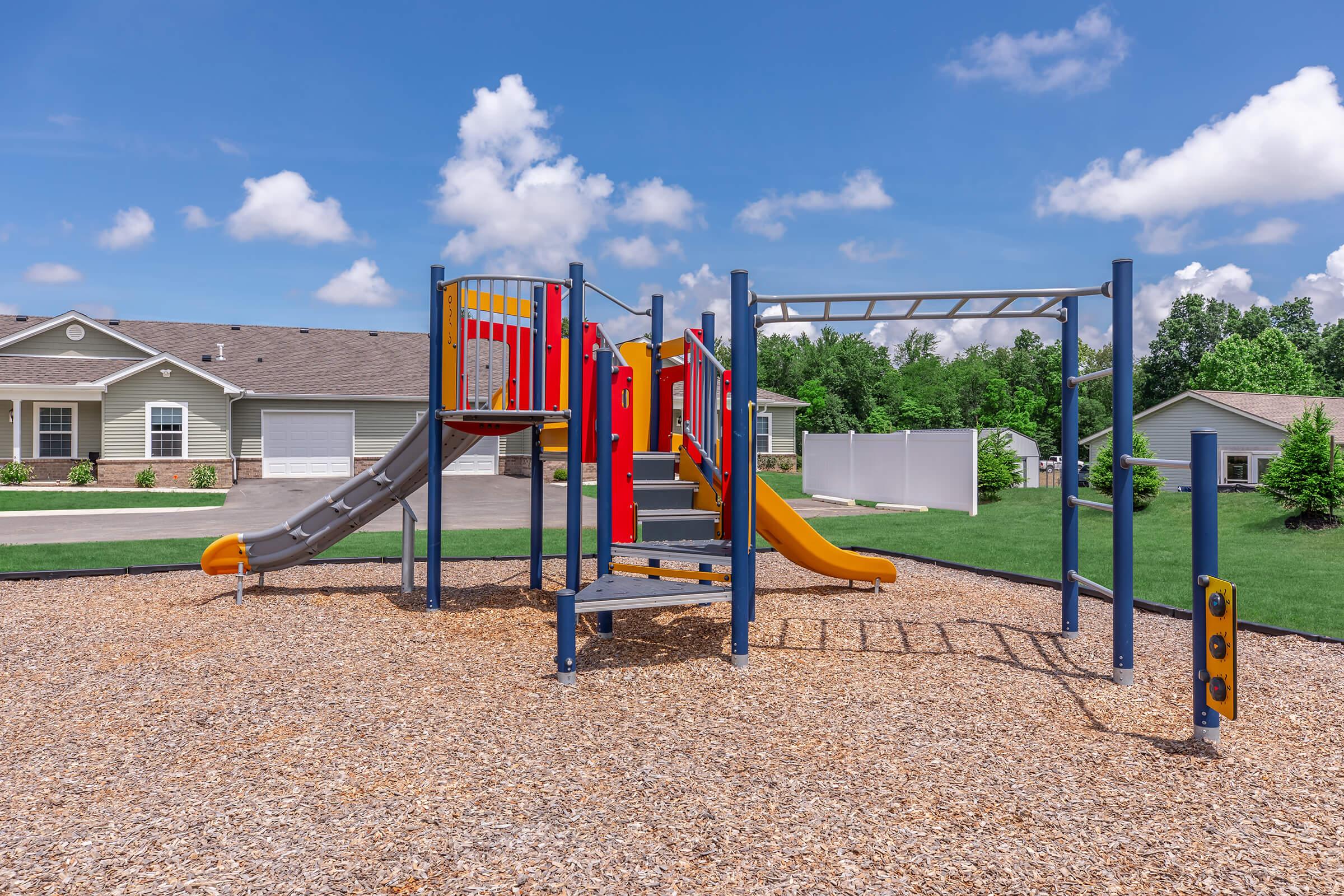 A colorful playground featuring slides, climbing structures, and a monkey bar, set in a sunny outdoor area with grassy surroundings and residential buildings in the background. Fluffy white clouds are visible in a blue sky, creating a cheerful atmosphere.