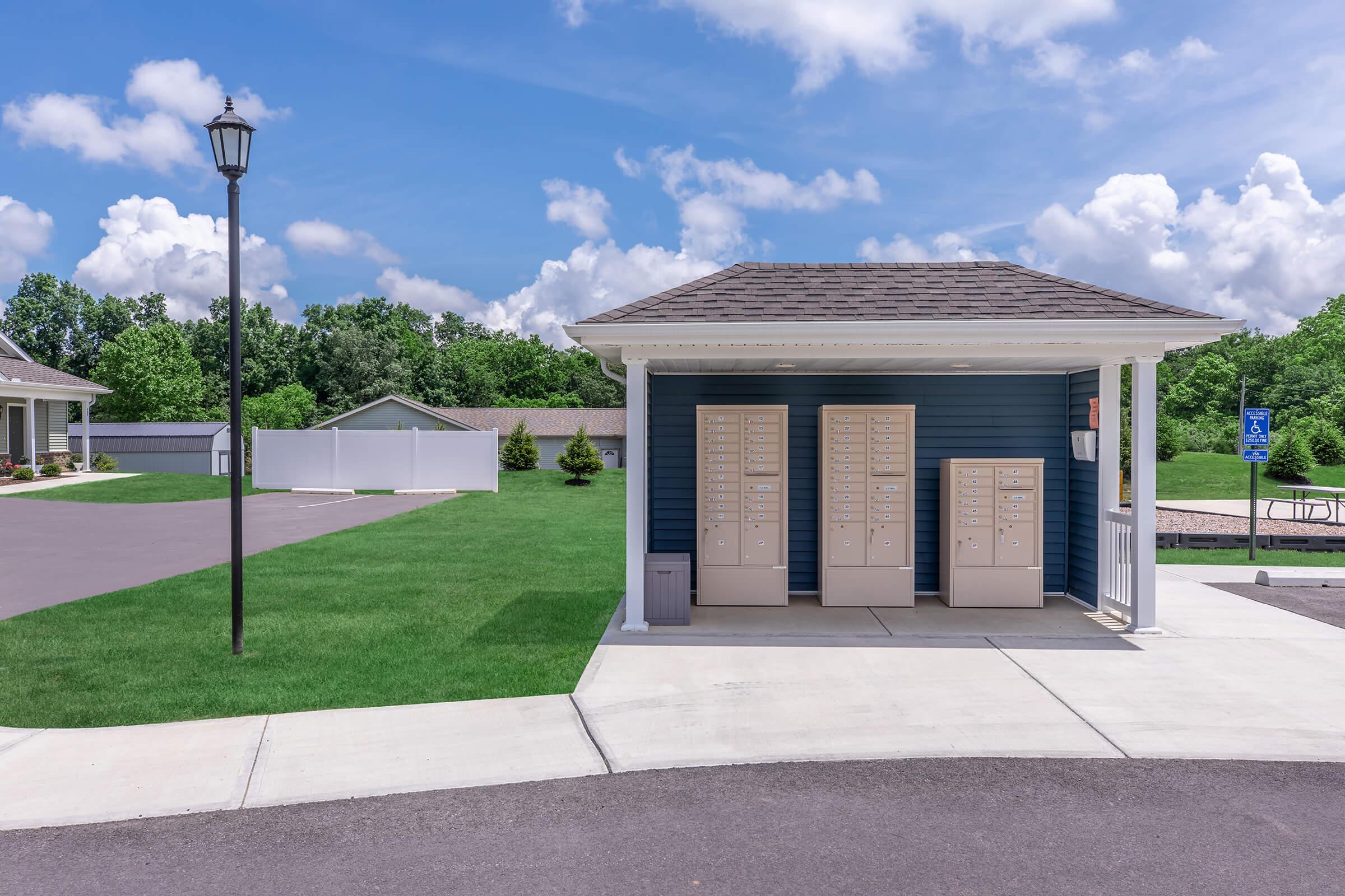 A small, blue building with a sloped roof housing beige mailboxes, surrounded by green grass and a paved walkway. A lamp post stands nearby, and in the background, there are trees and another building. The sky is bright with white clouds.