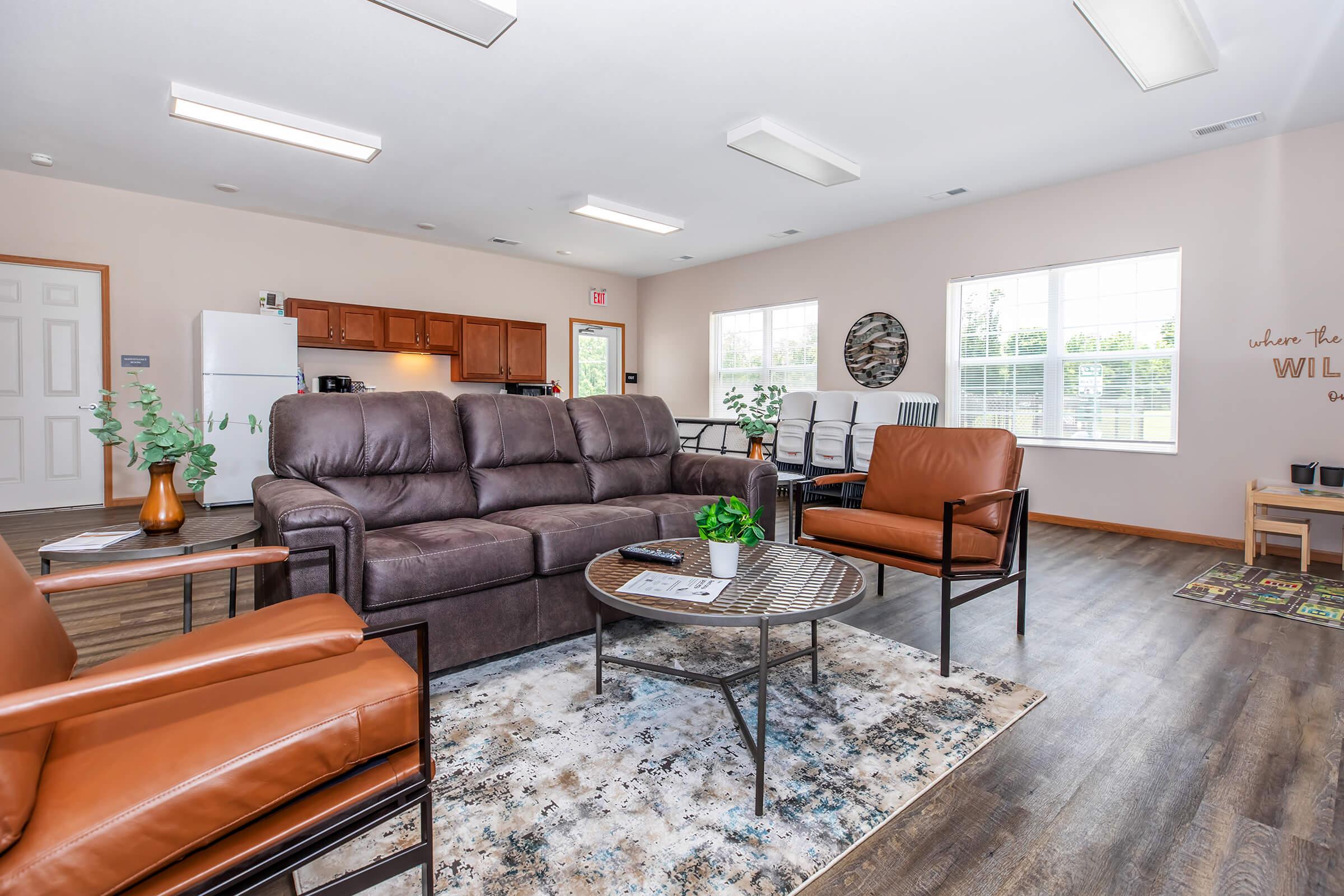 A cozy community lounge featuring a brown sectional sofa, two leather armchairs, a round coffee table with a small plant, and a colorful area rug. The space has warm lighting, a kitchenette in the background, and large windows allowing natural light. Decor includes a wall mural with motivational text.
