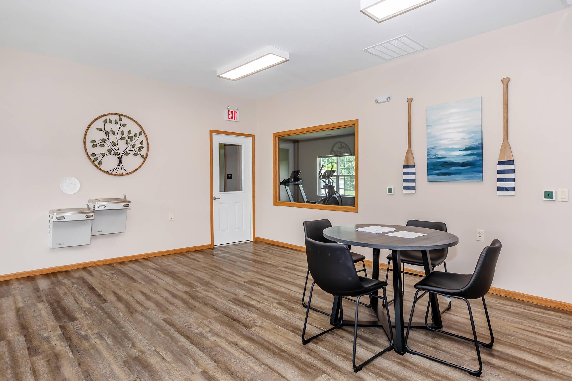 A well-lit room featuring a circular table with four black chairs. There are two mailboxes mounted on the wall, and a door leading to another area. A large window provides natural light, and decor includes a wall art piece with a tree design and a painting with blue tones. The floor has wooden planks.
