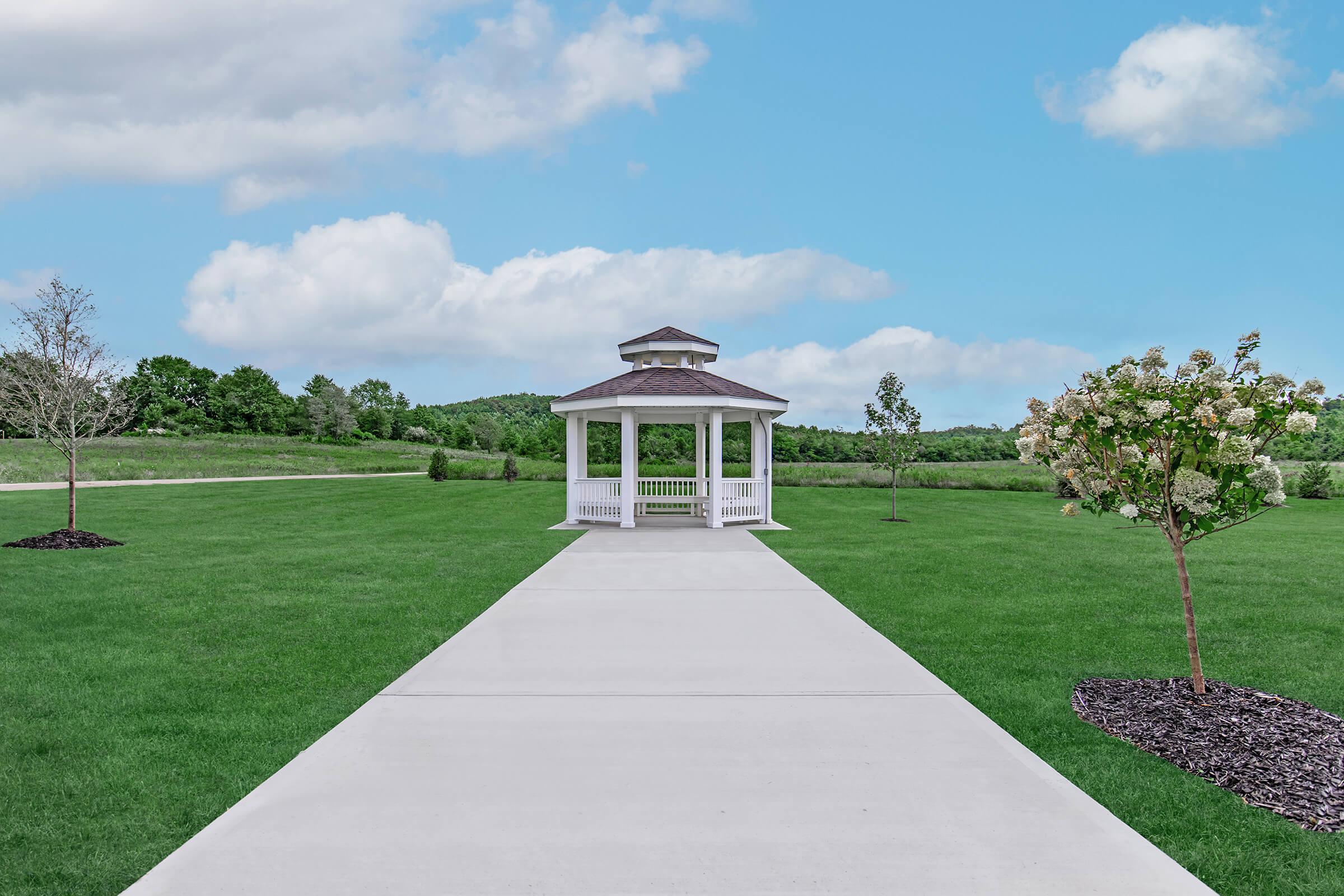 A white gazebo with a peaked roof sits at the end of a concrete pathway in a lush green landscape. The pathway is flanked by manicured grass and small trees, with a blue sky and fluffy white clouds in the background, creating a serene outdoor setting.