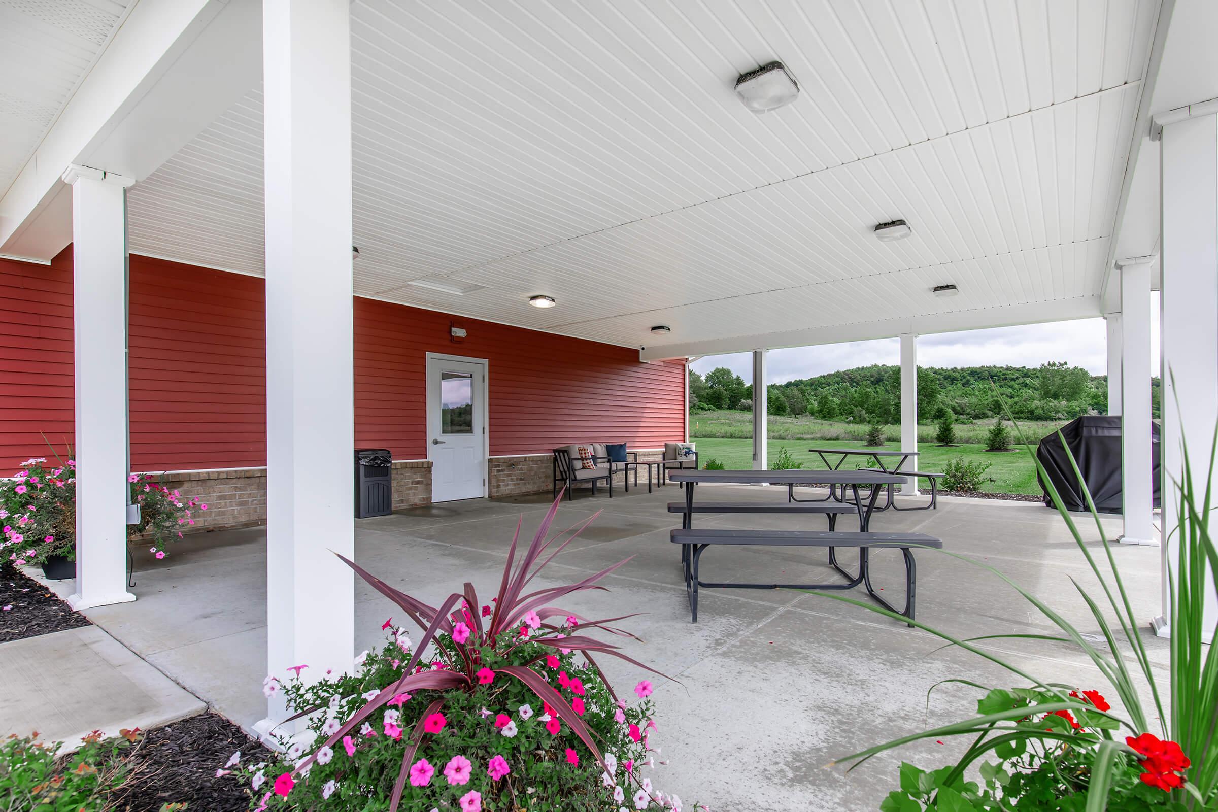 An outdoor covered area with benches and tables, surrounded by colorful flowers. The structure has a red exterior wall and white support pillars. A grassy landscape is visible in the background under a partly cloudy sky.