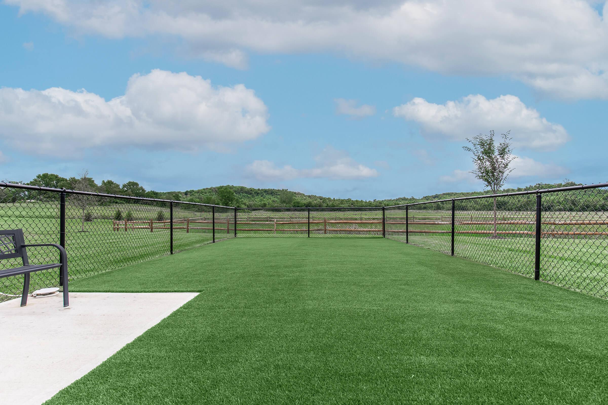 A spacious, fenced-in area with artificial grass, featuring a concrete section on the left and a bench. The background showcases lush green hills under a partly cloudy blue sky, creating a serene outdoor environment.