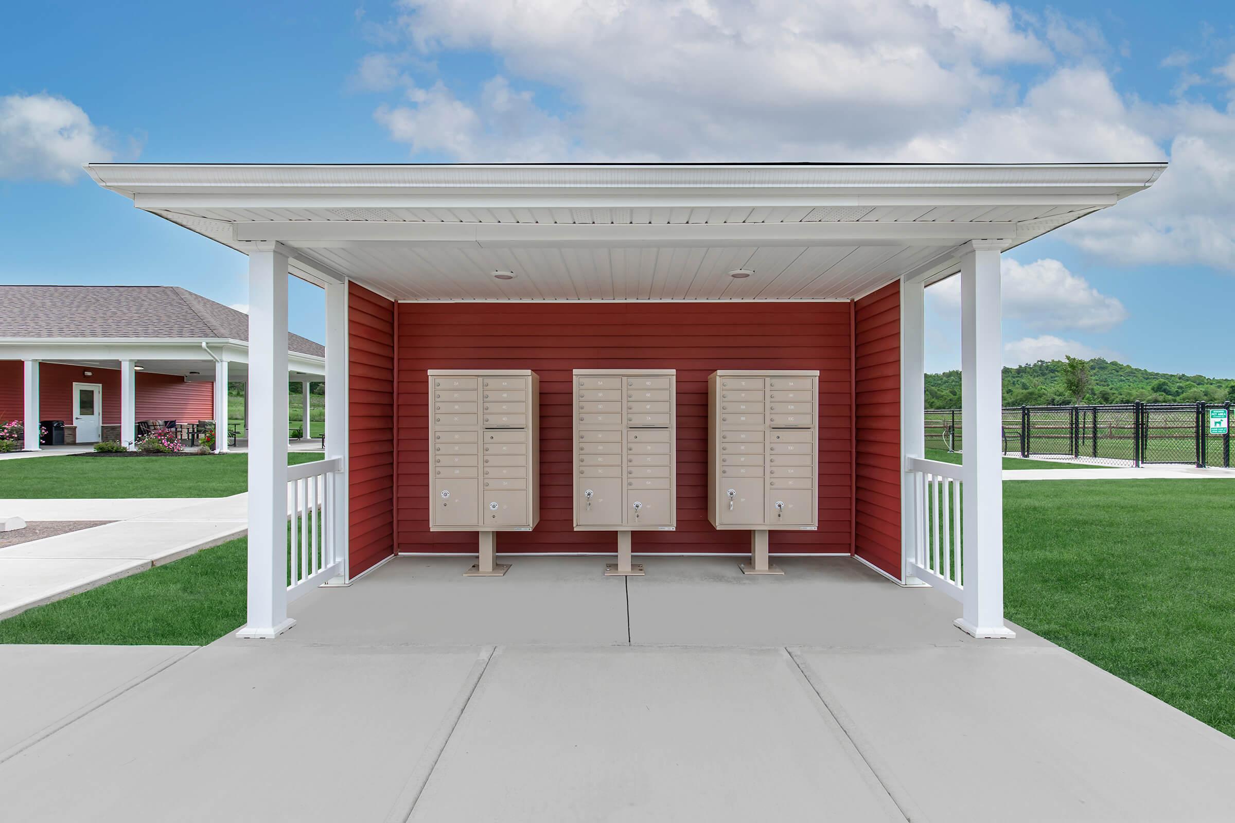 A covered outdoor area with three beige mailboxes mounted on posts, set against a red building. The surrounding area features green grass and a concrete walkway, with a clear blue sky in the background. The setting appears well-maintained and spacious.