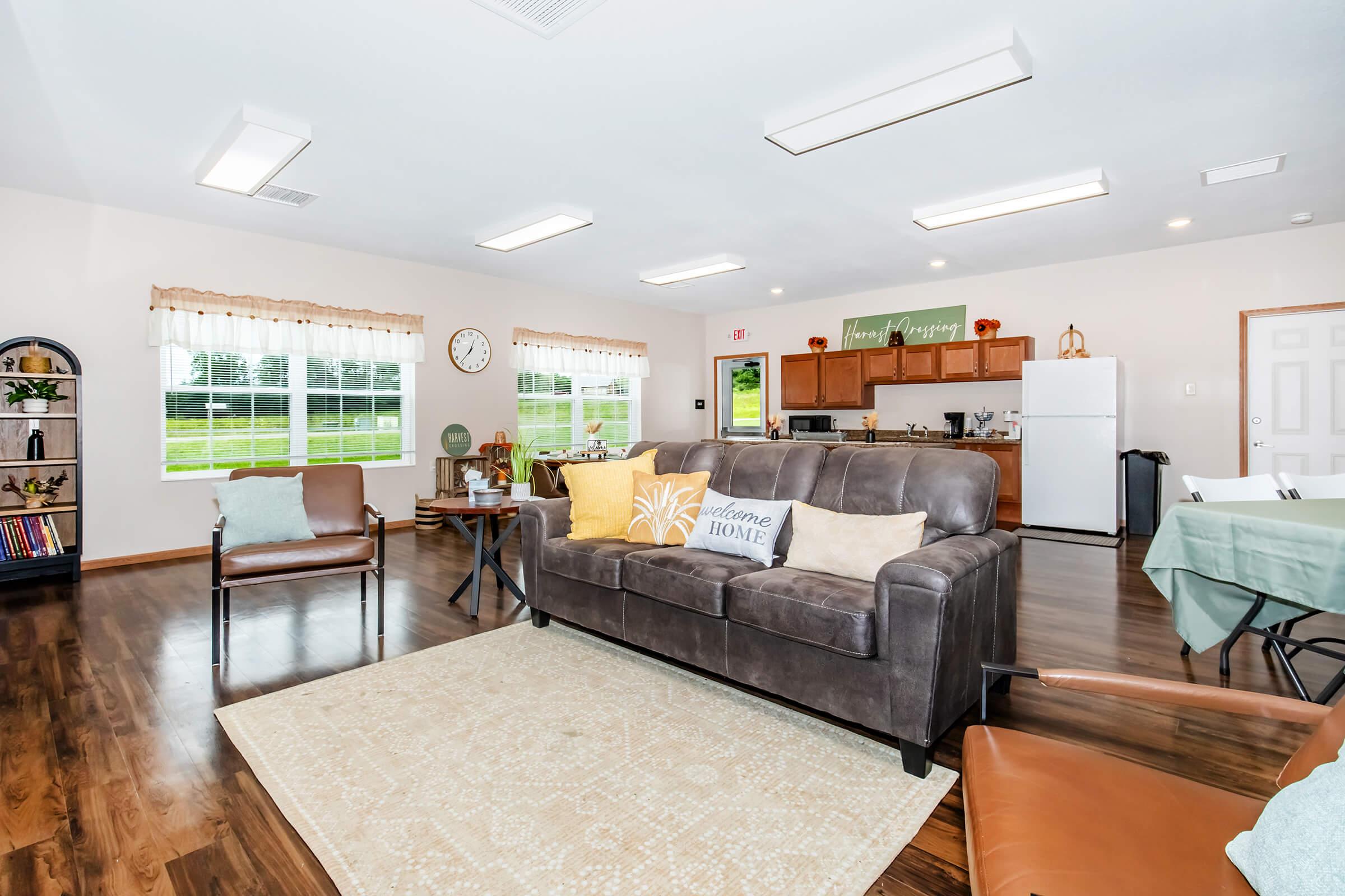 A cozy common room featuring a comfortable brown couch with decorative pillows, a light-colored rug, and a wooden table with chairs. The space includes a kitchenette with cabinets, a refrigerator, and a clock on the wall. Large windows provide ample natural light, and there are decorative elements that enhance the welcoming atmosphere.