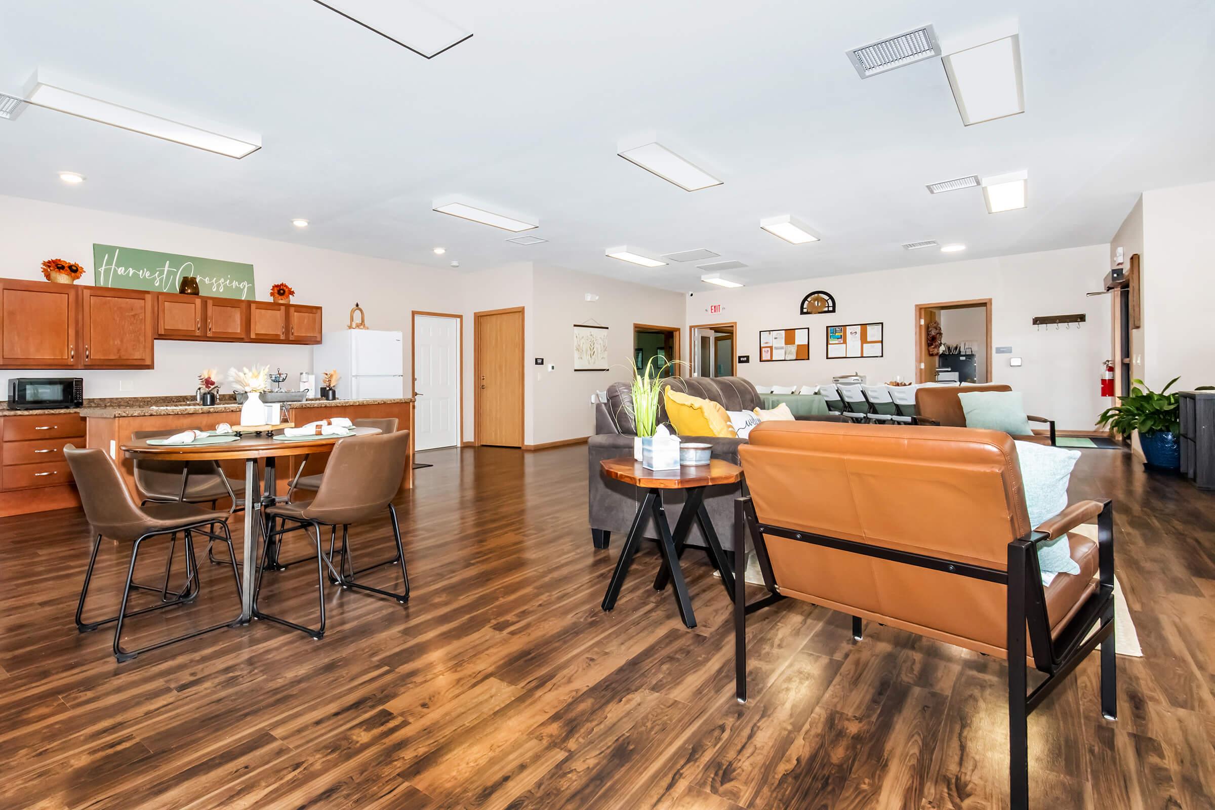 A spacious, modern common area featuring a dining table with chairs, a cozy seating arrangement with brown and teal sofas, and a kitchenette with wooden cabinets. The floor is dark wood, and there are bright overhead lights. Walls are light-colored with a decorative sign saying "Homemade Goodness."