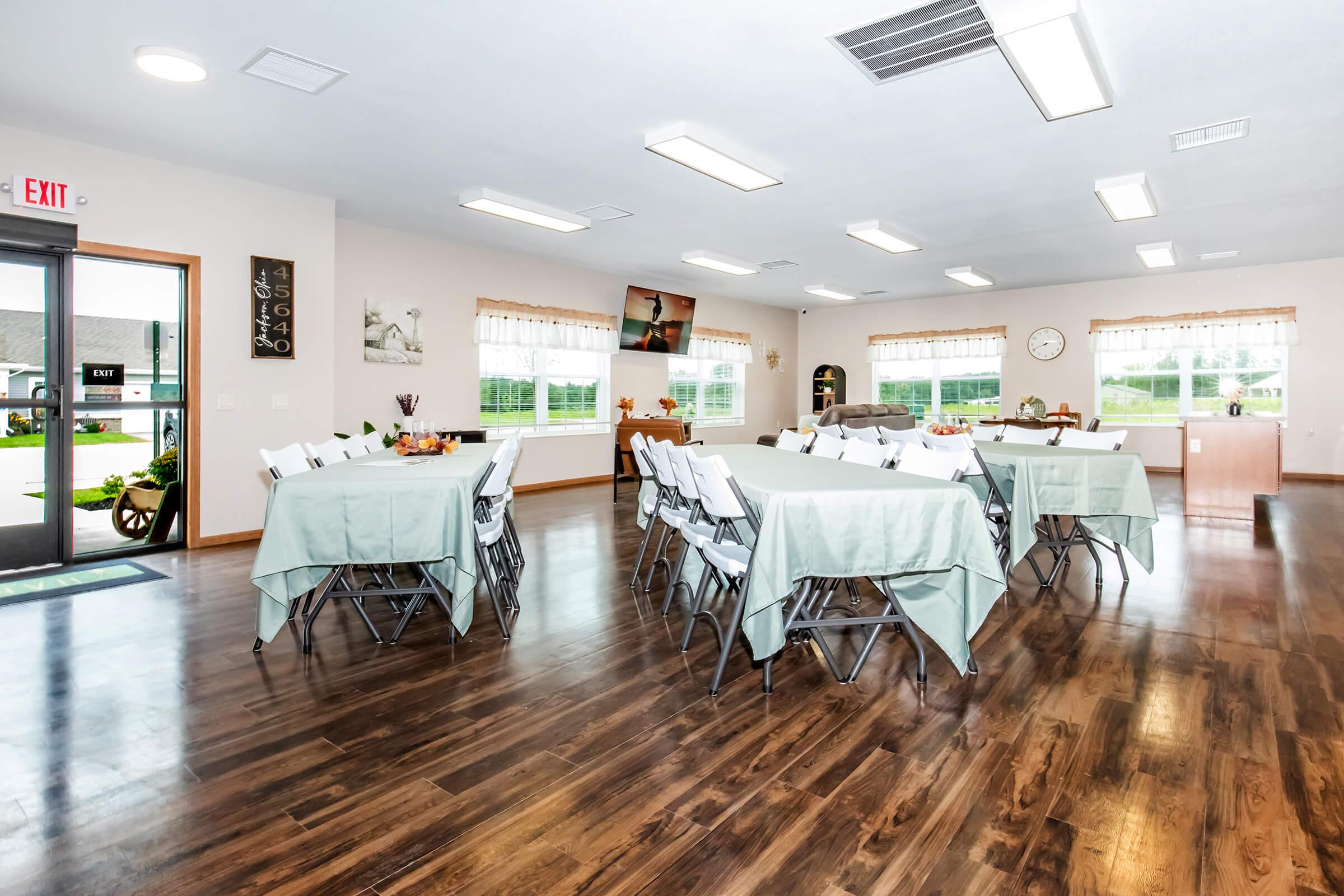 A spacious, well-lit room set up for events, featuring multiple tables covered with light blue tablecloths and white chairs. The décor includes a wall clock, framed pictures, and large windows providing natural light. An exit door is visible, along with a television displaying a scenic image.
