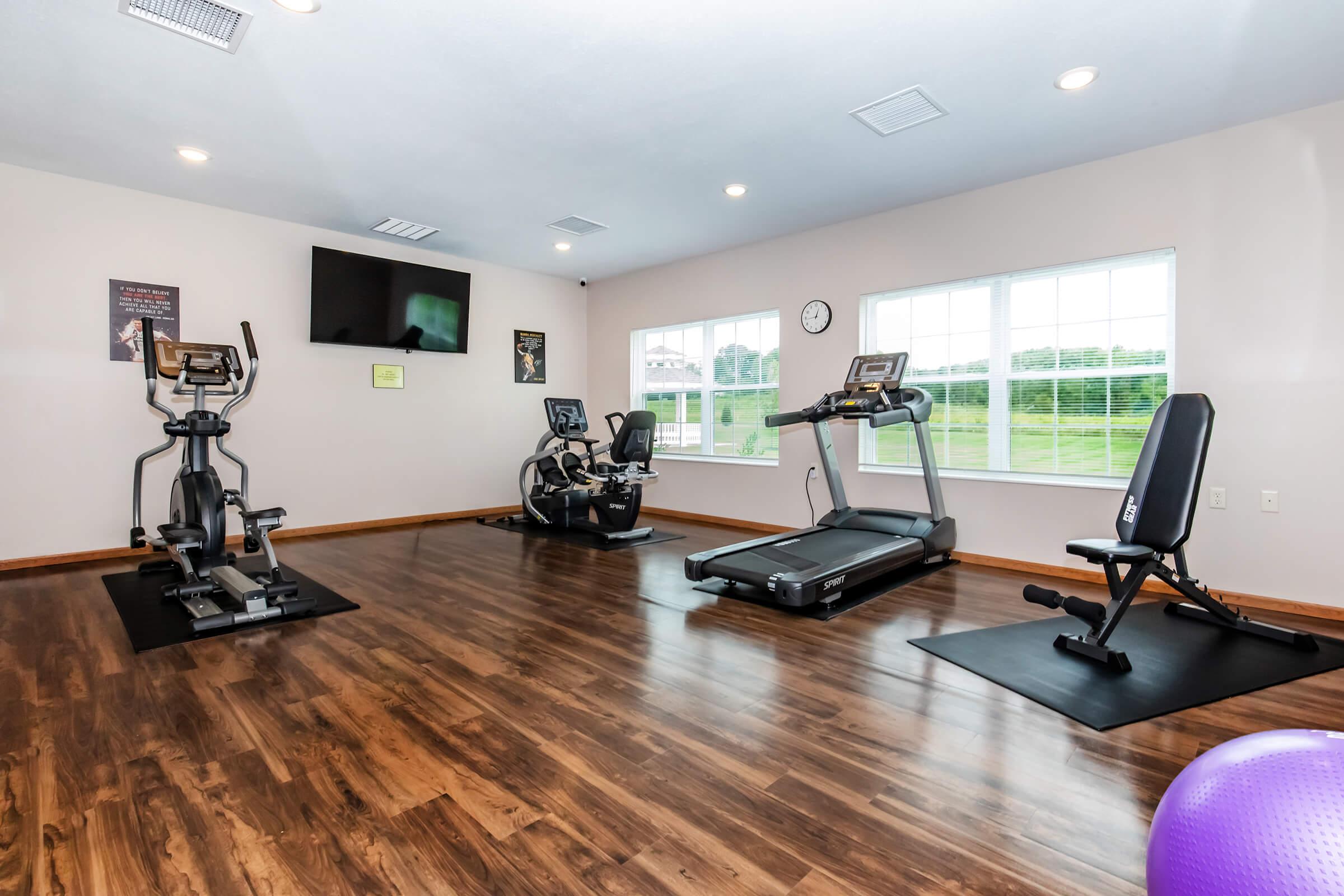 A modern fitness room featuring various exercise equipment, including an elliptical machine, a treadmill, and a weight bench. Large windows allow natural light, and there’s a clock on the wall. The floor is wooden, and a large TV is mounted, creating an inviting workout environment.