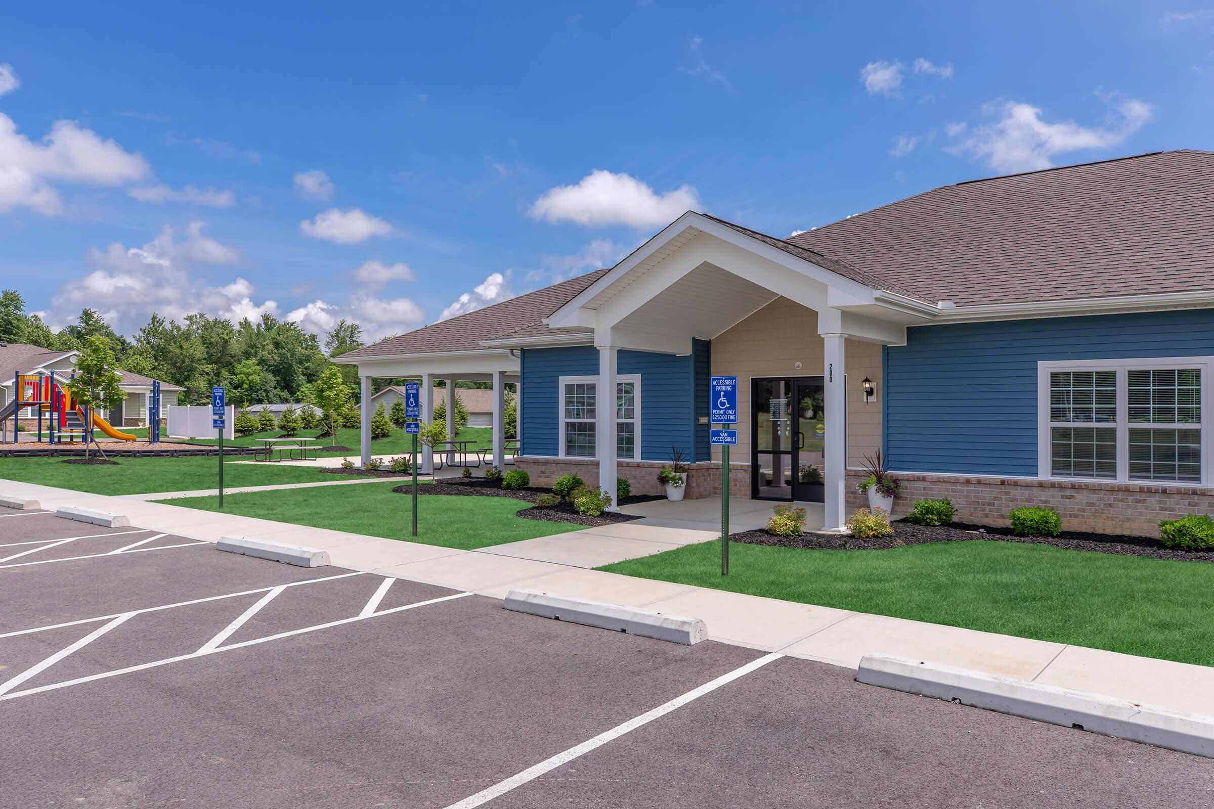 A modern blue building with a welcoming entrance, surrounded by well-maintained landscaping and a spacious parking lot. Nearby, a playground is visible, providing recreational space. The sky is clear with scattered clouds, enhancing the bright, inviting atmosphere of the area.