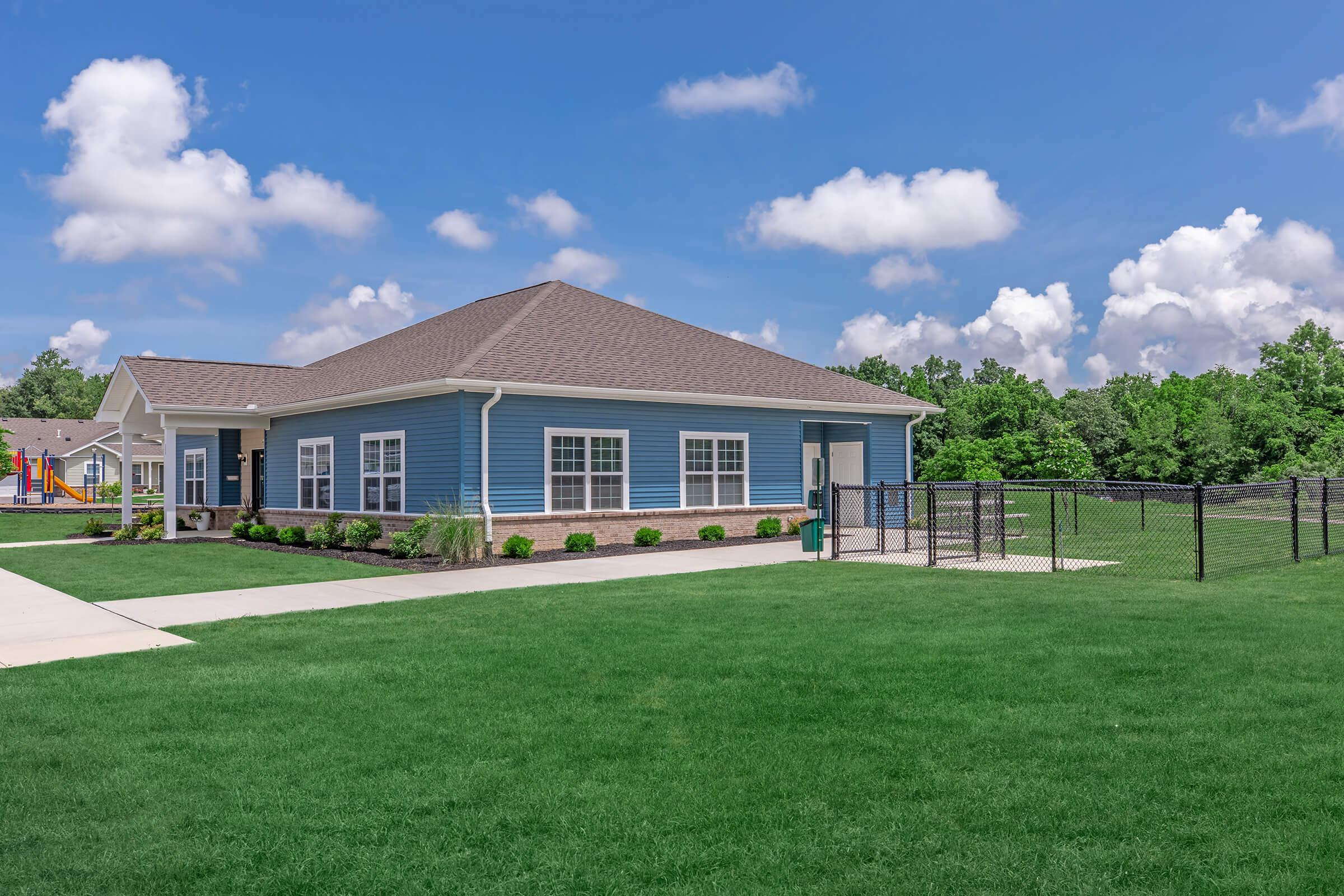 A modern blue house with a brown shingled roof is situated on a well-maintained green lawn. The front features a concrete walkway and flower beds, with a playground visible in the background. White fluffy clouds float in a bright blue sky, and a fenced area is visible beside the house.
