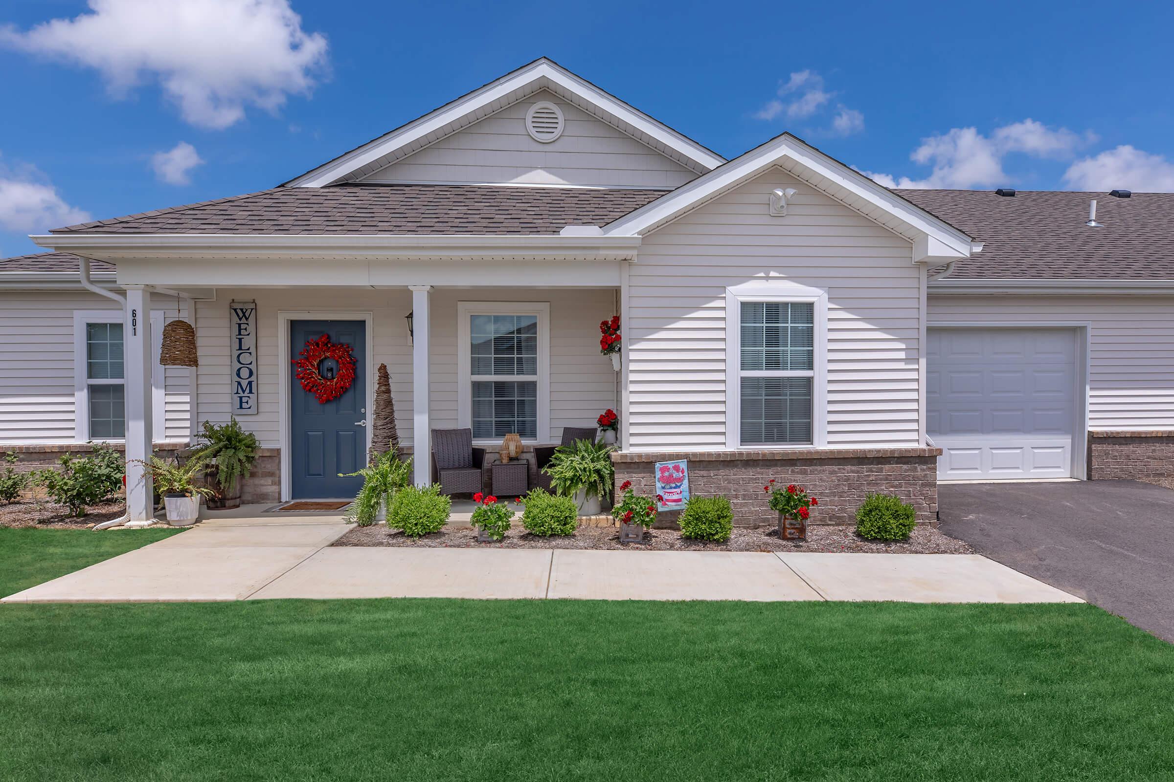A well-maintained single-story house with a welcoming front porch. The entrance features a blue door with a heart decoration, surrounded by potted plants and flowers. A neatly manicured lawn and driveway lead to a garage on the right. The sky is bright with fluffy clouds.