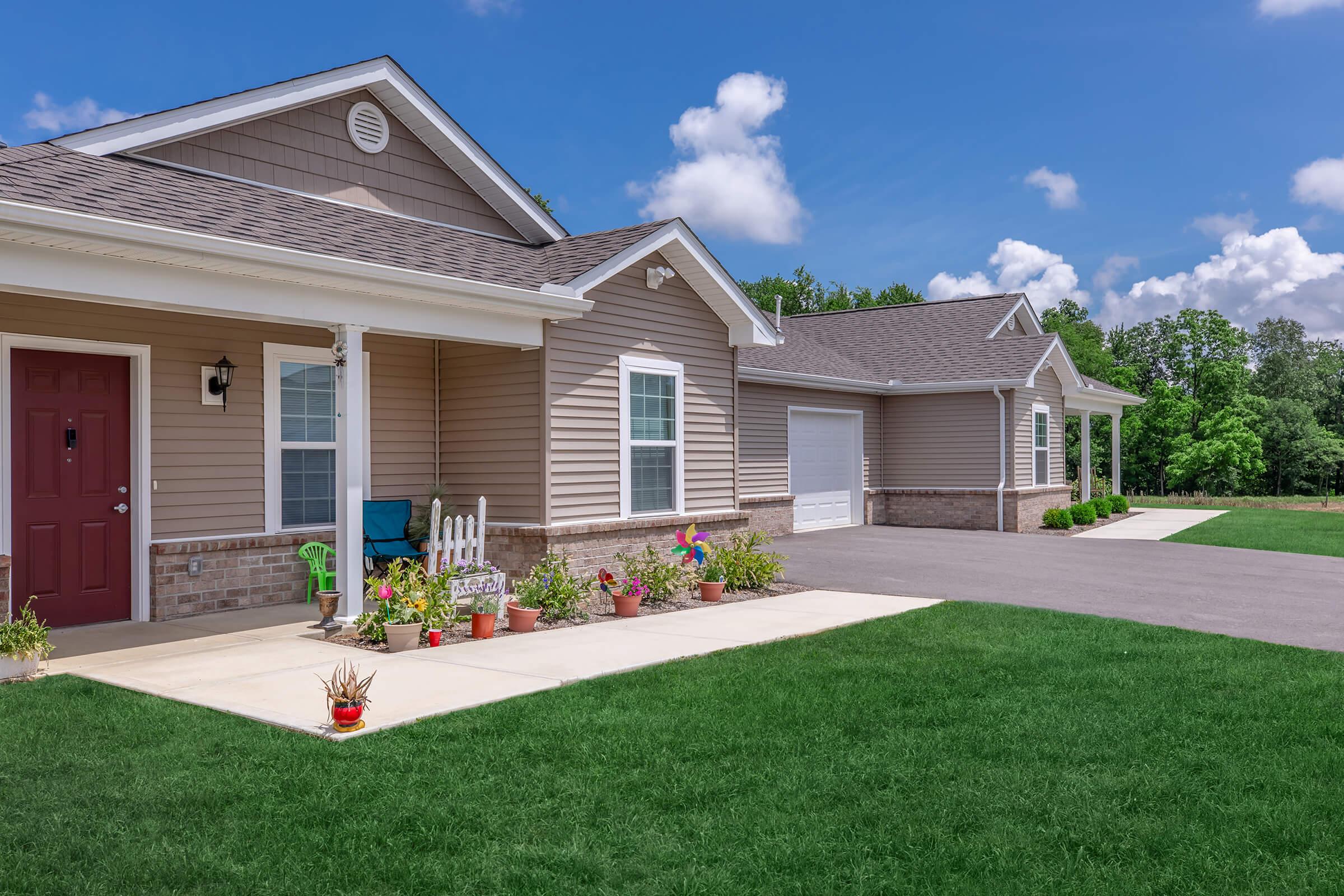 A well-maintained single-story home with a porch, surrounded by a green lawn and various potted plants. The driveway leads to a garage on the side, under a clear blue sky with a few clouds, creating a serene suburban atmosphere.