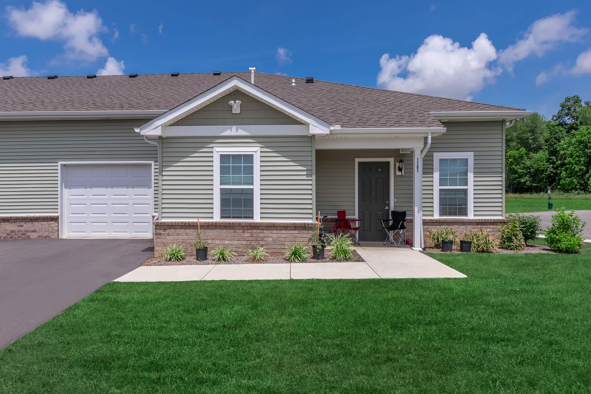 A single-story house featuring a light gray exterior with a garage, front porch, and landscaping. The driveway is paved, and there are potted plants along the walkway leading to the entrance. The sky is bright blue with a few clouds, and there is greenery in the background.