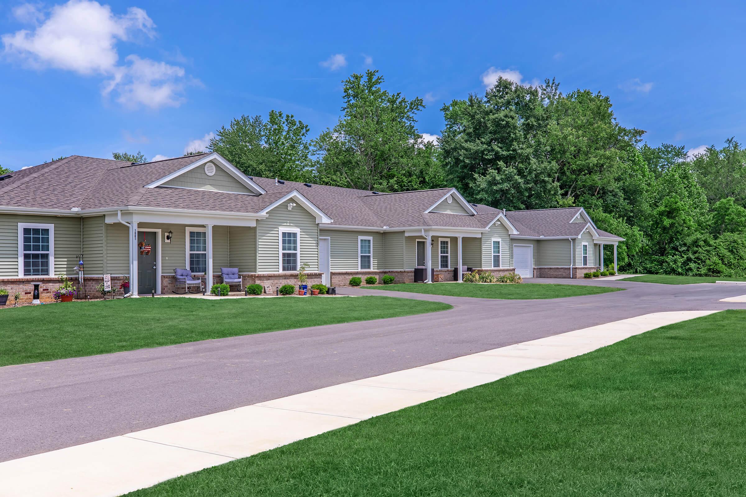 A row of single-story houses with light gray siding, situated along a paved driveway. Each house features a porch with seating and landscaping, including green grass and flower beds. The background includes dense trees under a clear blue sky with some clouds.