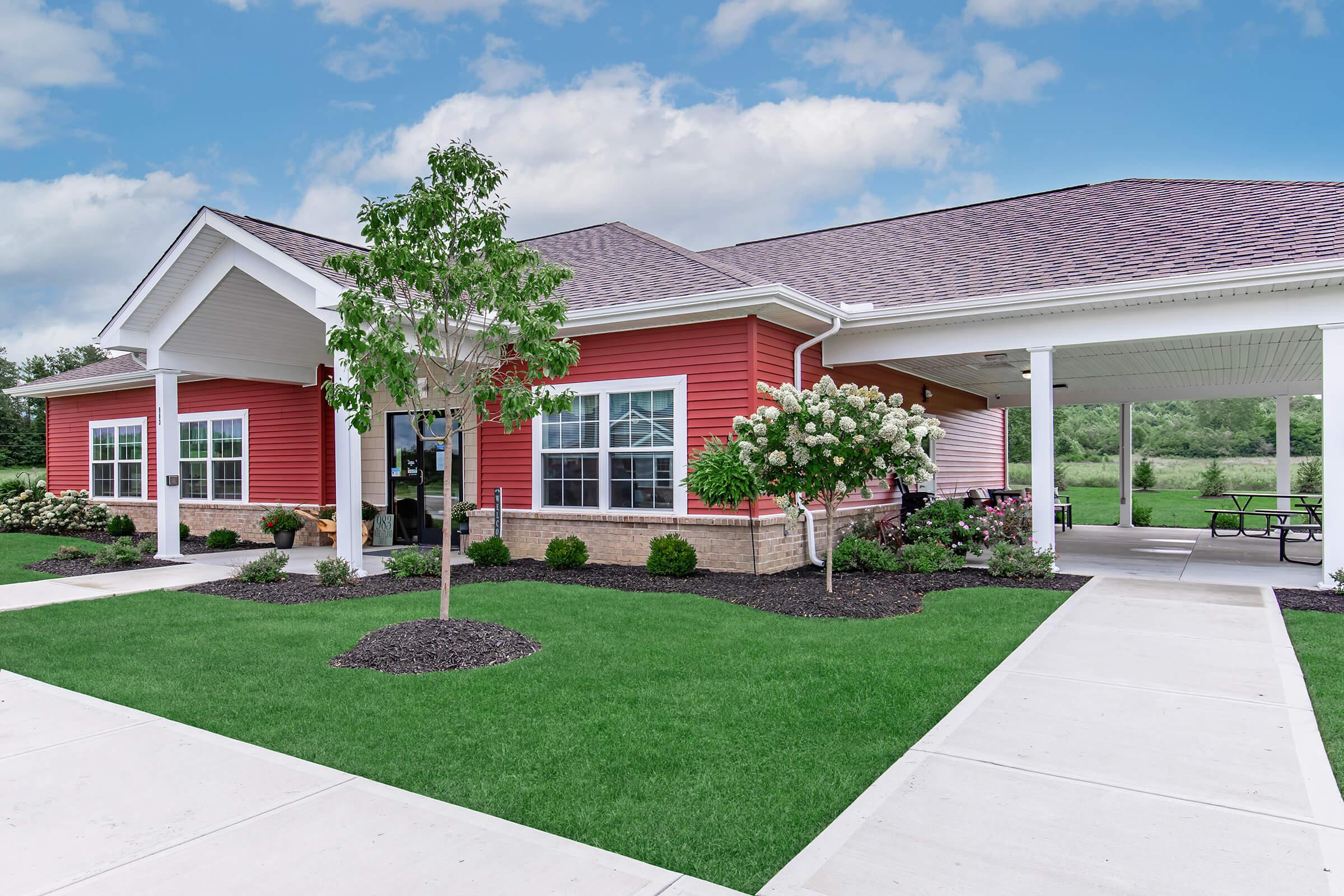 A vibrant red building with a welcoming entrance, surrounded by well-maintained green lawns and blooming shrubs. The structure features a sloped roof and large windows, with a covered outdoor area and picnic tables visible nearby. The scene is set against a backdrop of blue skies and fluffy clouds.