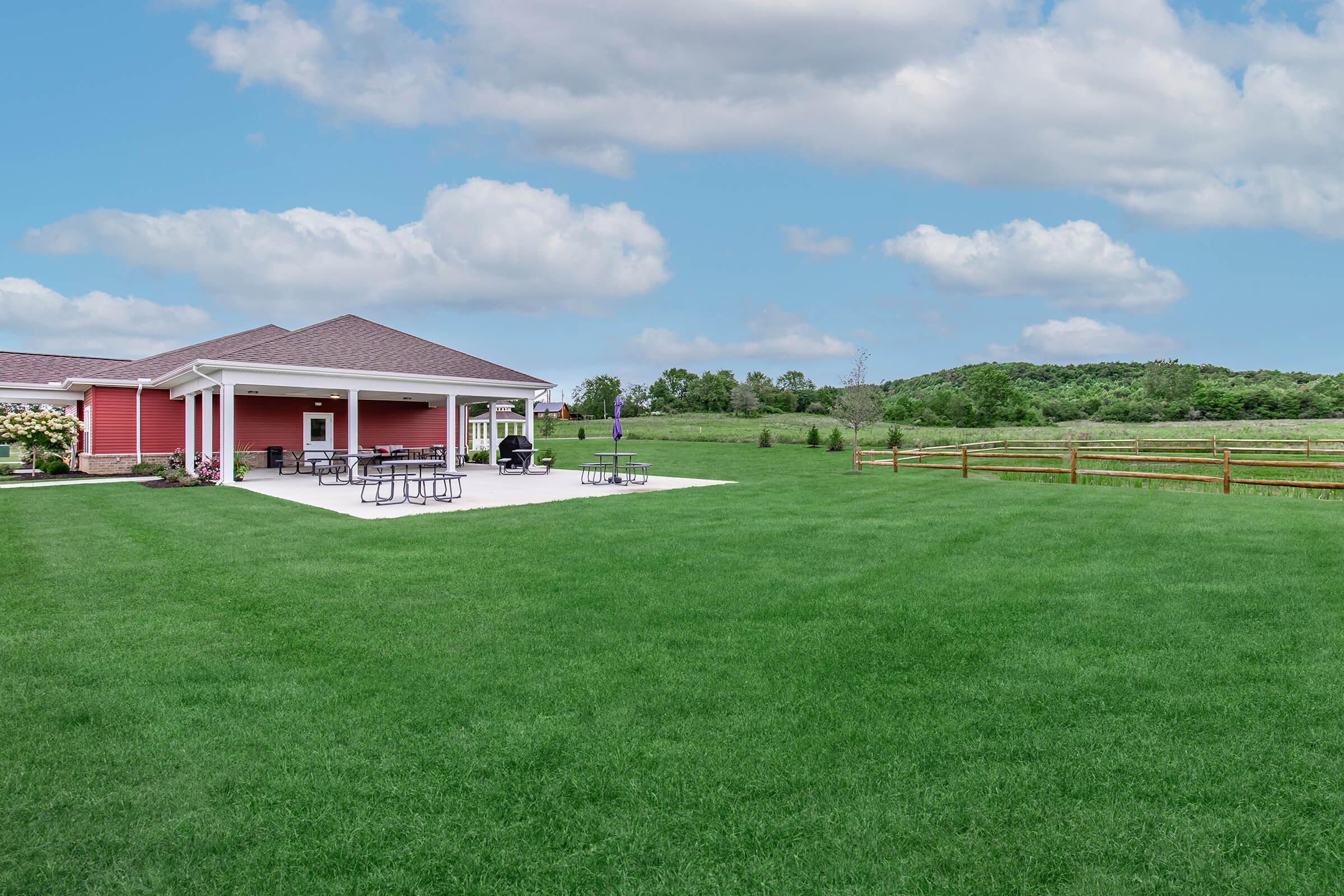 A spacious lawn with green grass in front of a red building featuring a covered patio. Several picnic tables are arranged on the patio, and a wooden fence borders the area. In the background, rolling hills and a blue sky with fluffy clouds create a serene landscape.