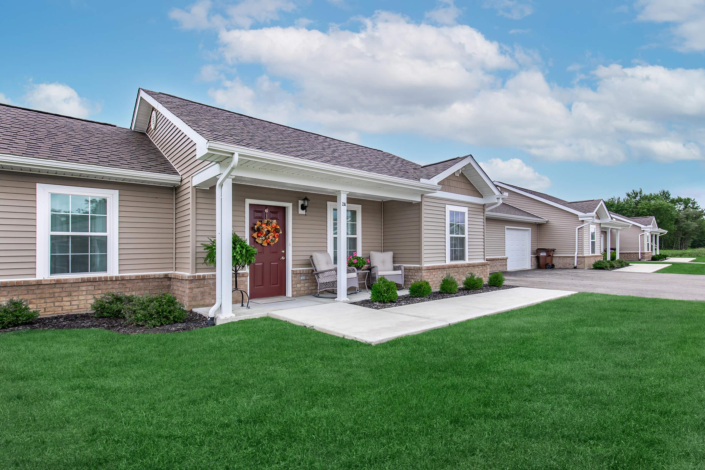 A well-maintained single-story home with a light brown exterior, featuring a covered porch with chairs and a decorative wreath on the door. The front yard is lush and green, bordered by small plants. In the background, there are similar homes along a paved driveway under a blue sky with scattered clouds.