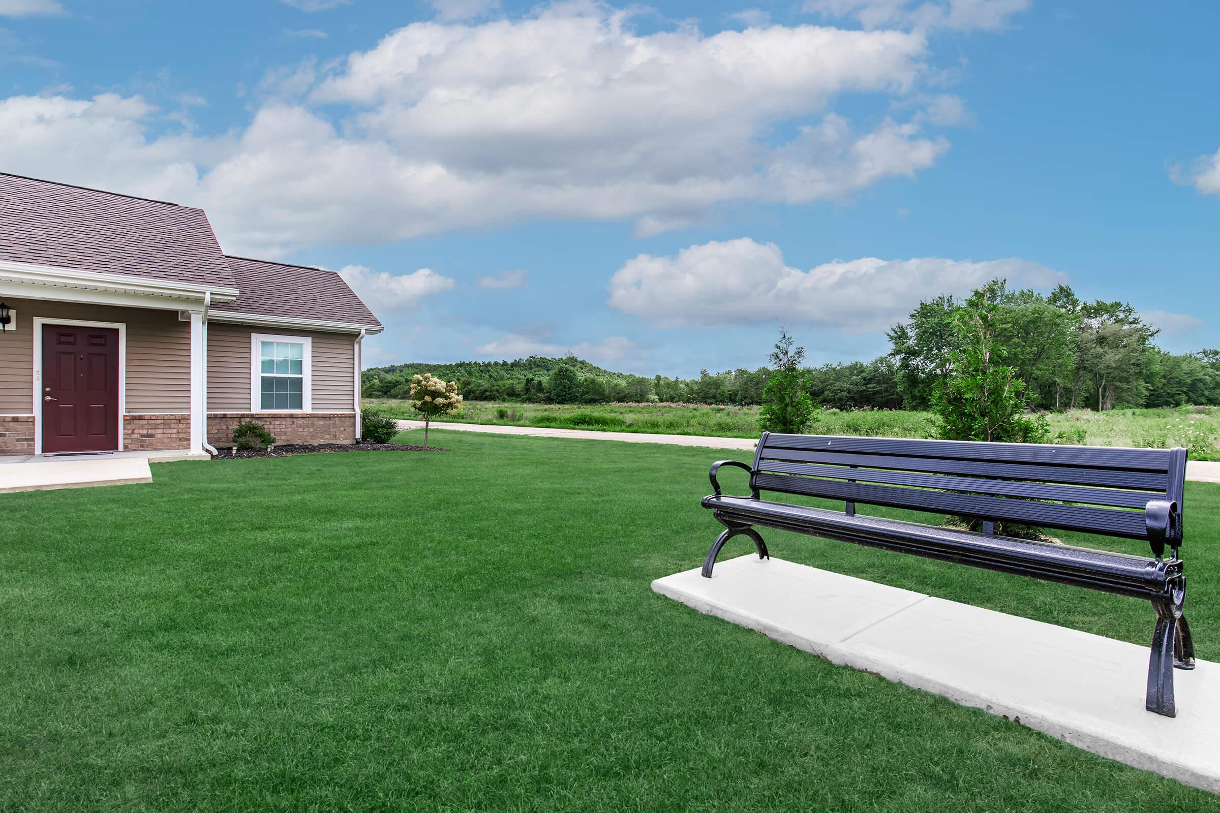 A well-maintained yard featuring a green lawn and a black bench positioned on a concrete pathway. In the background, a single-story house with a brown exterior and a reddish roof is visible, along with lush trees and open fields under a partly cloudy sky.