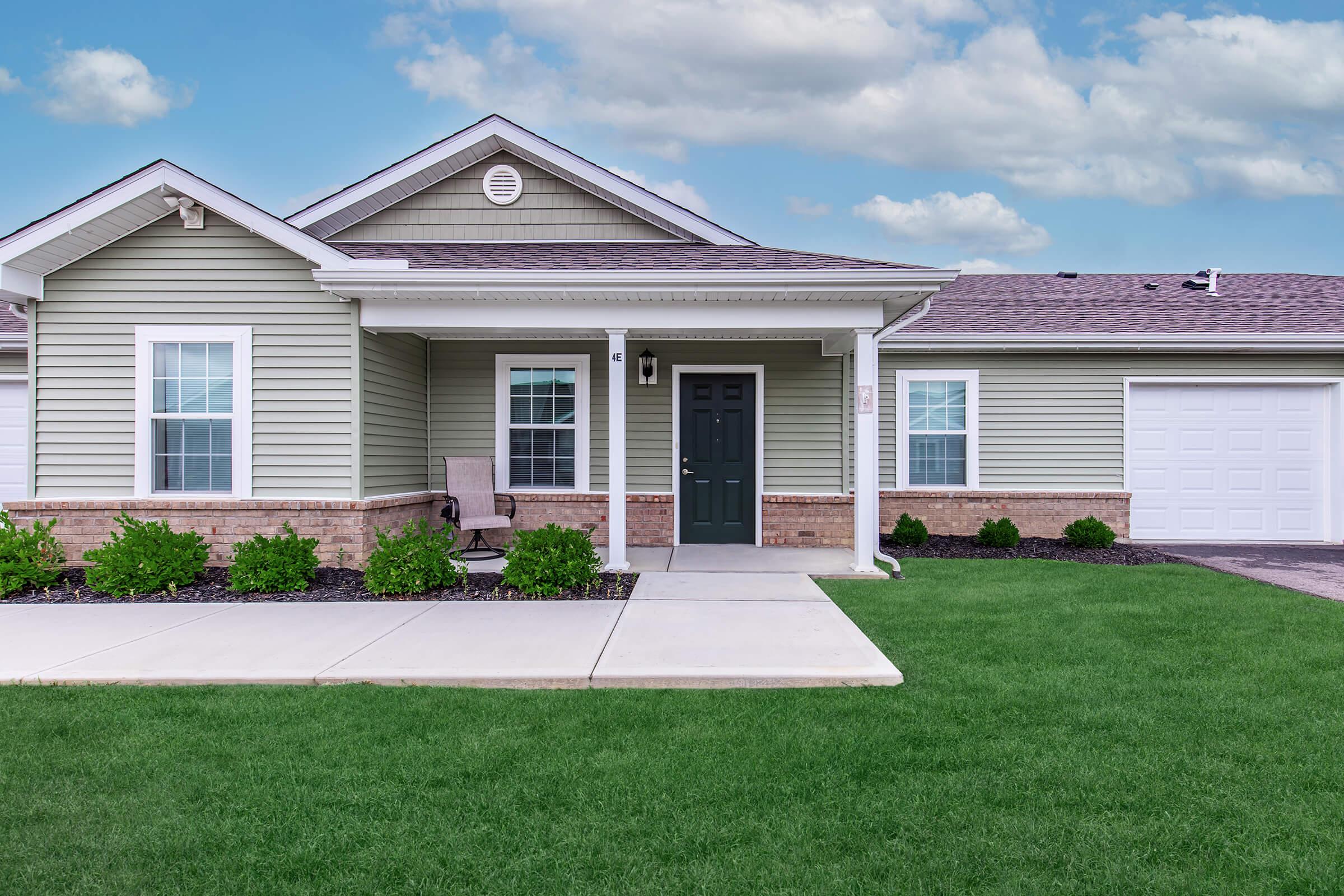 A single-story home with light green siding and a dark front door. The porch features a chair and is flanked by small shrubs. The lawn is well-maintained, and there is a garage on the right side of the house. The sky is partly cloudy, adding a pleasant backdrop to the scene.