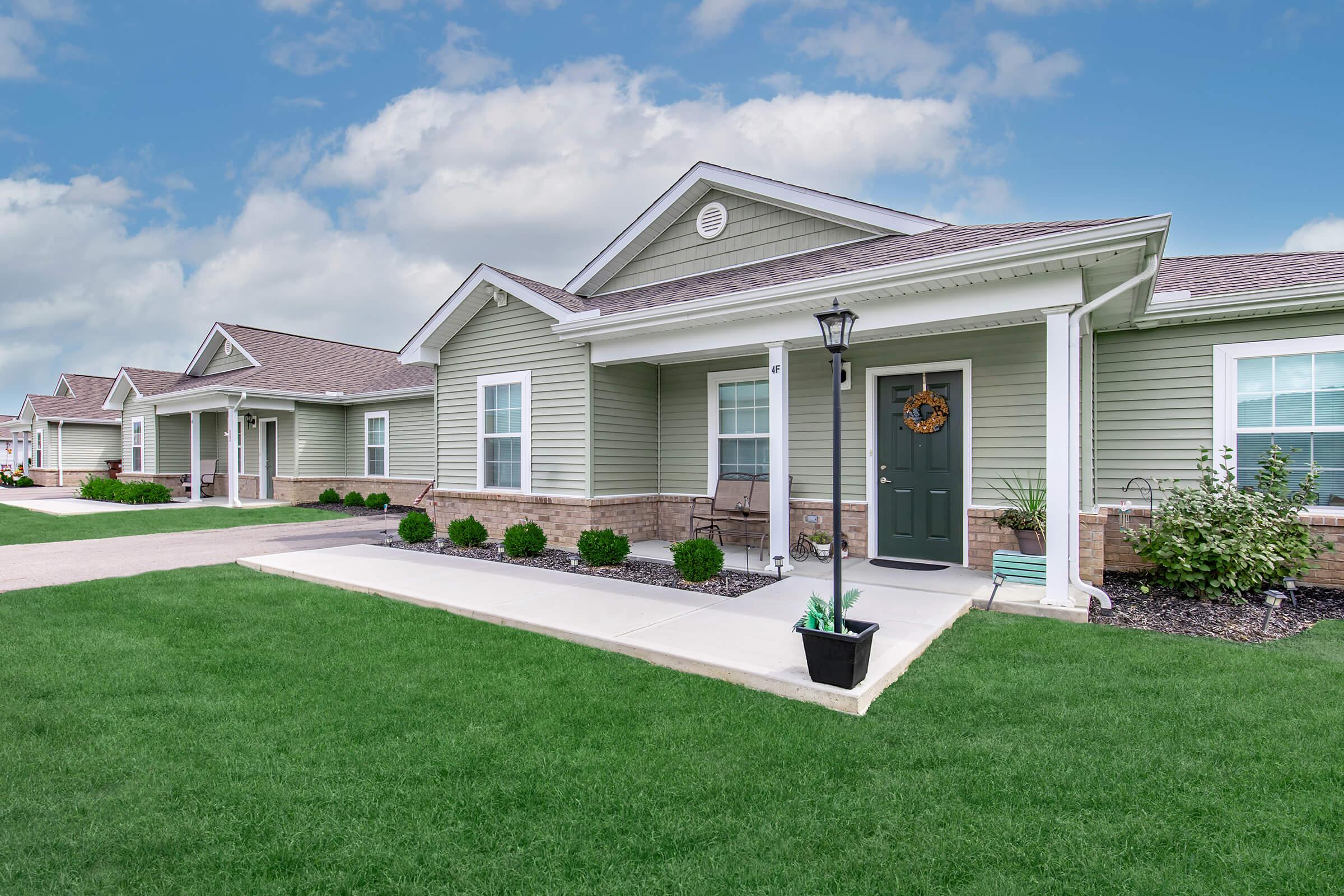 Single-story residential building with a green exterior and a welcoming front porch. The walkway leads to a green door adorned with a wreath. Lush green lawn and neatly trimmed shrubs are visible, along with additional houses in the distance under a partly cloudy sky.