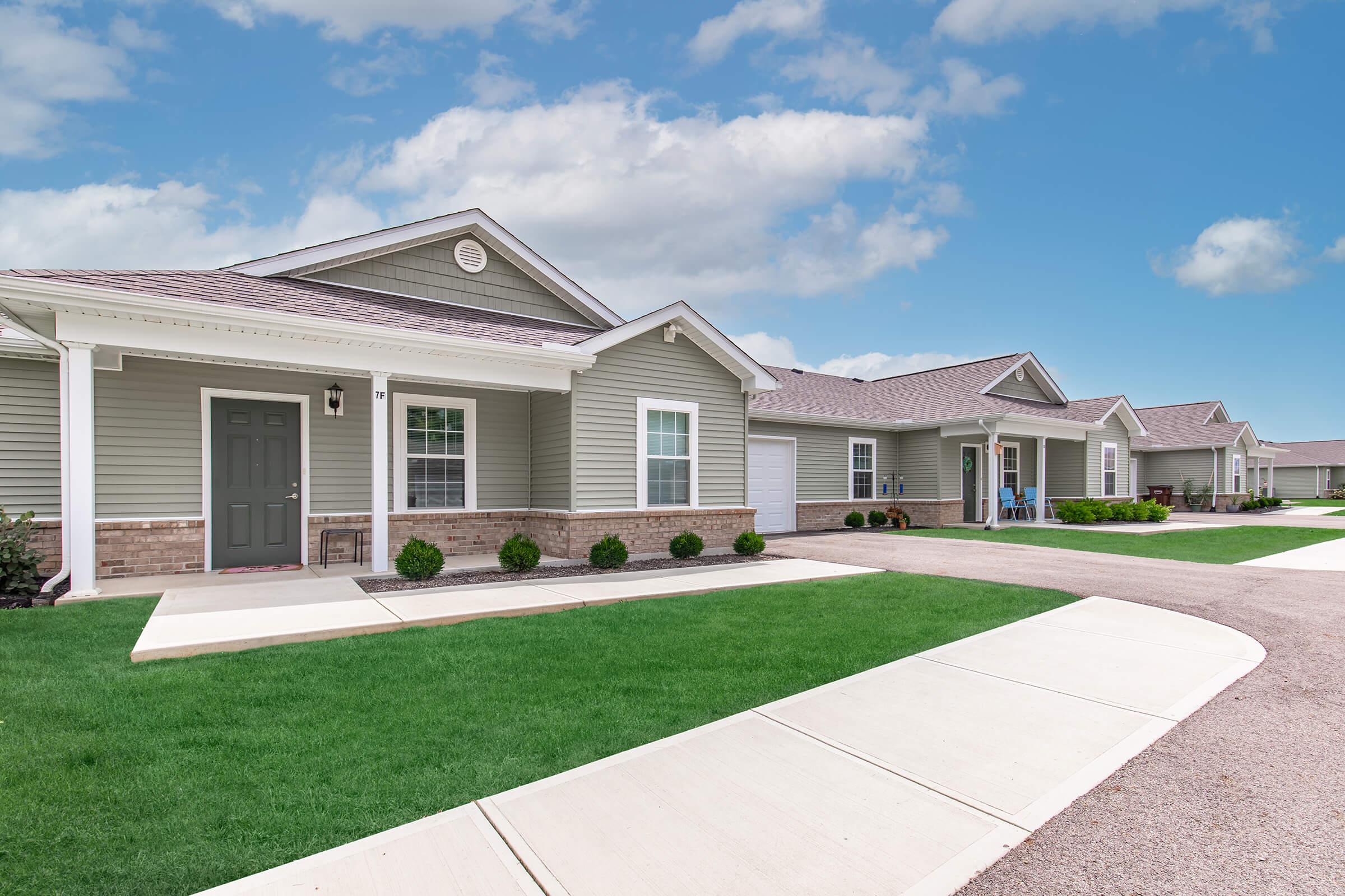 A row of modern single-family homes with light gray siding and brick accents. Each house features a front porch, green lawns, and a concrete walkway. The sky is partly cloudy, creating a bright atmosphere. The setting appears well-maintained and inviting.