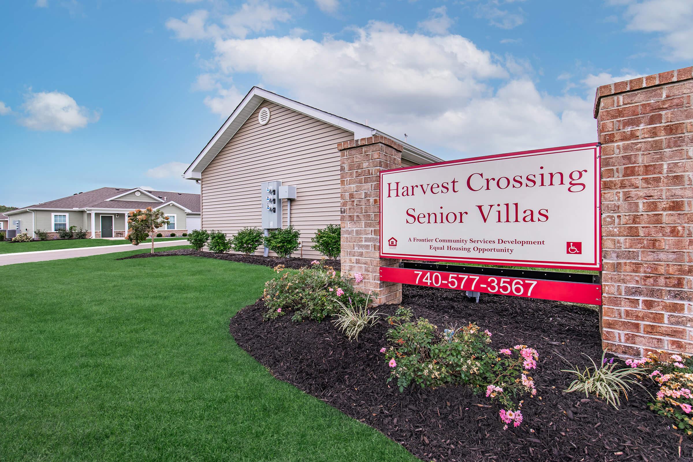 Sign for Harvest Crossing Senior Villas in a landscaped area, featuring well-maintained grass and flower beds. The building in the background is a single-story residential structure. The sign indicates the property focuses on providing housing for seniors and includes contact information.