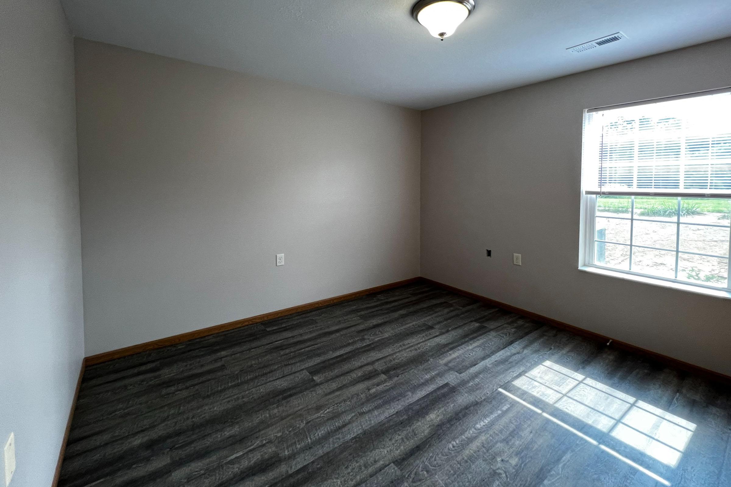 A well-lit, empty room with light gray walls and a brown wooden floor. A single window allows natural light to enter, and there is a ceiling light fixture. The room appears freshly painted and ready for furniture.
