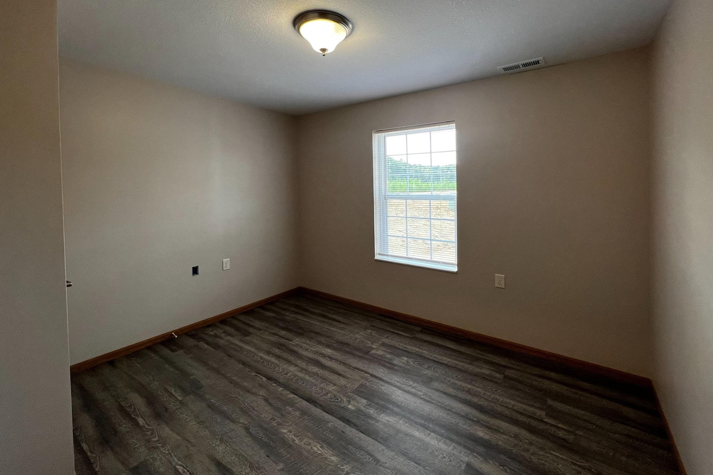 A vacant room with light brown walls and a wooden floor. There is a single window allowing natural light and a ceiling light fixture. The room appears empty, with no furniture or decorations.
