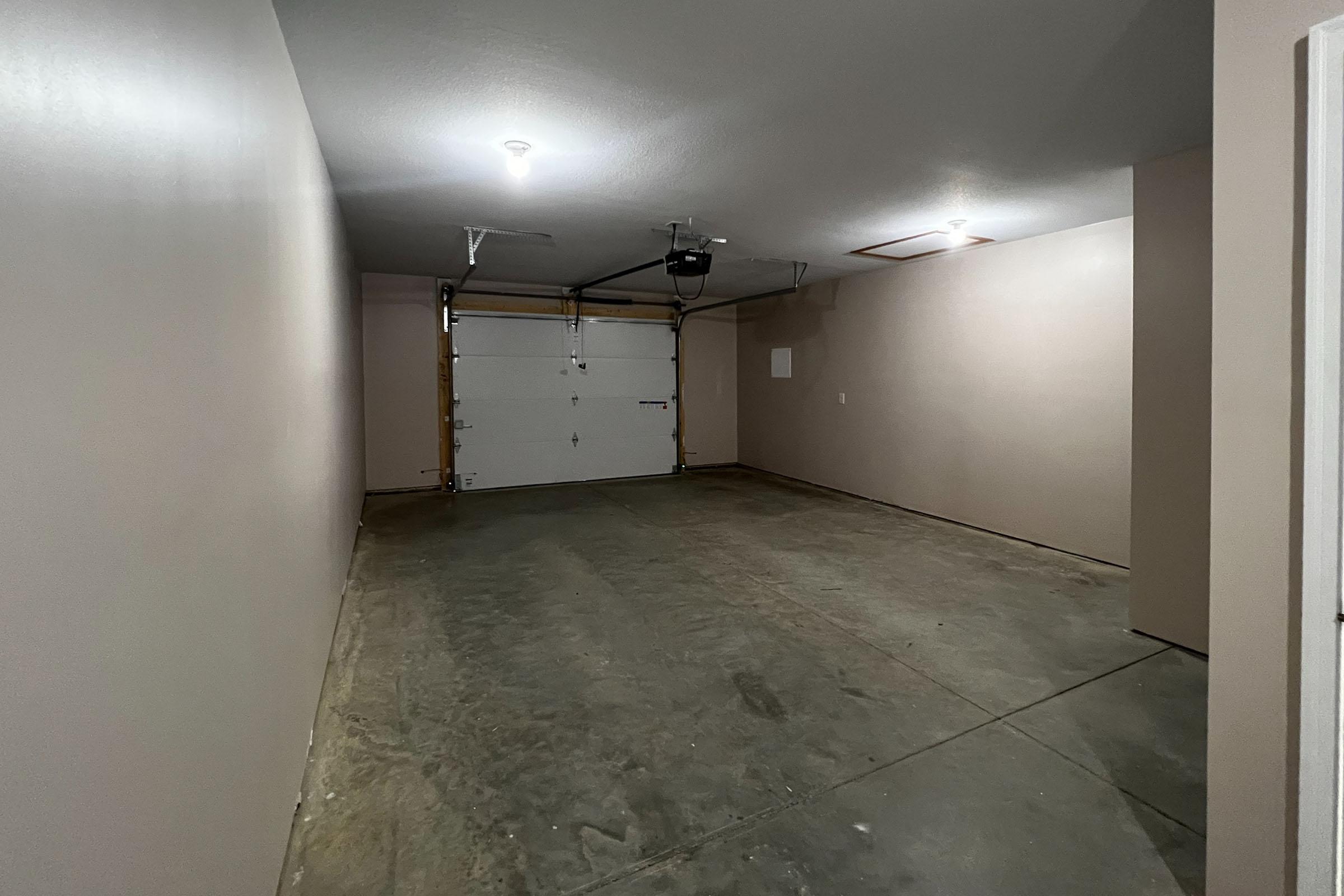 Empty garage with a smooth concrete floor, featuring a single white garage door and plain beige walls. The space is well-lit by overhead lighting, and there are no vehicles or items present, creating a clean and open environment.