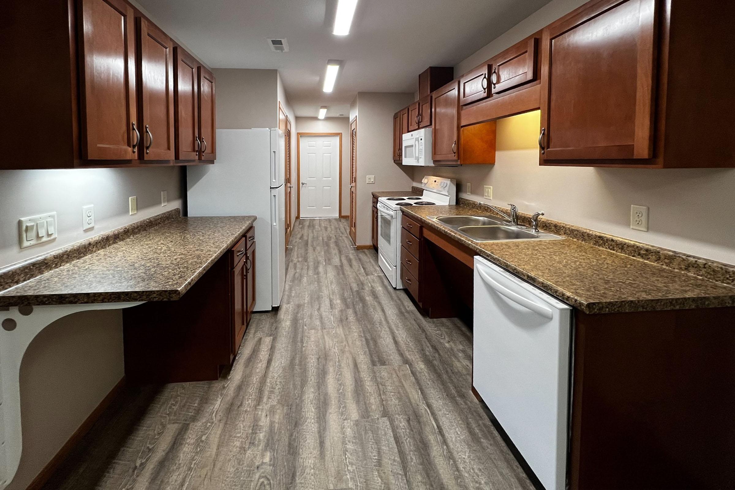 A modern kitchen featuring wooden cabinets, a light-colored countertop, and a white refrigerator. The kitchen includes an oven and stove, a sink, and ample storage space. The flooring is a light wood laminate, and the space is well-lit with overhead lighting. A door at the back leads to another area.