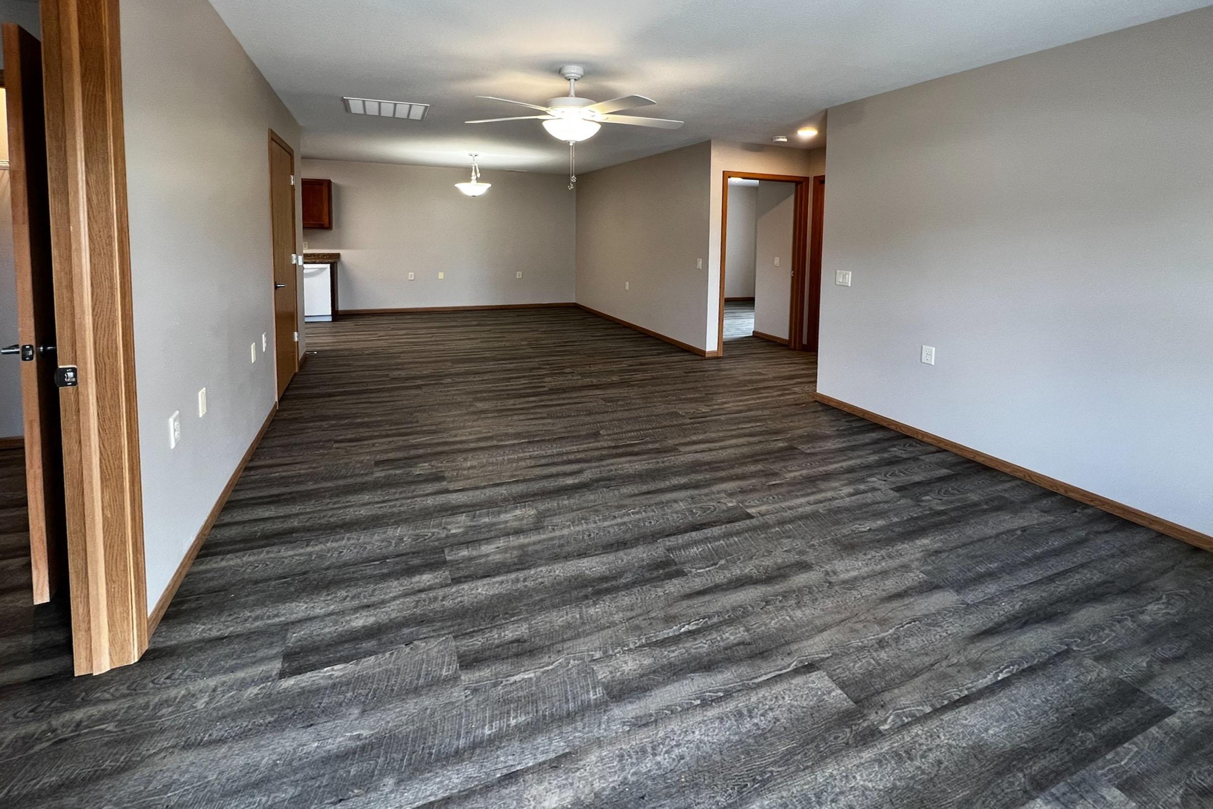 Spacious interior of a modern living area featuring dark laminate flooring, neutral-colored walls, and wooden door frames. There are ceiling fans above, with doorways leading to other rooms. Natural light illuminates the space, giving it an open and inviting atmosphere.