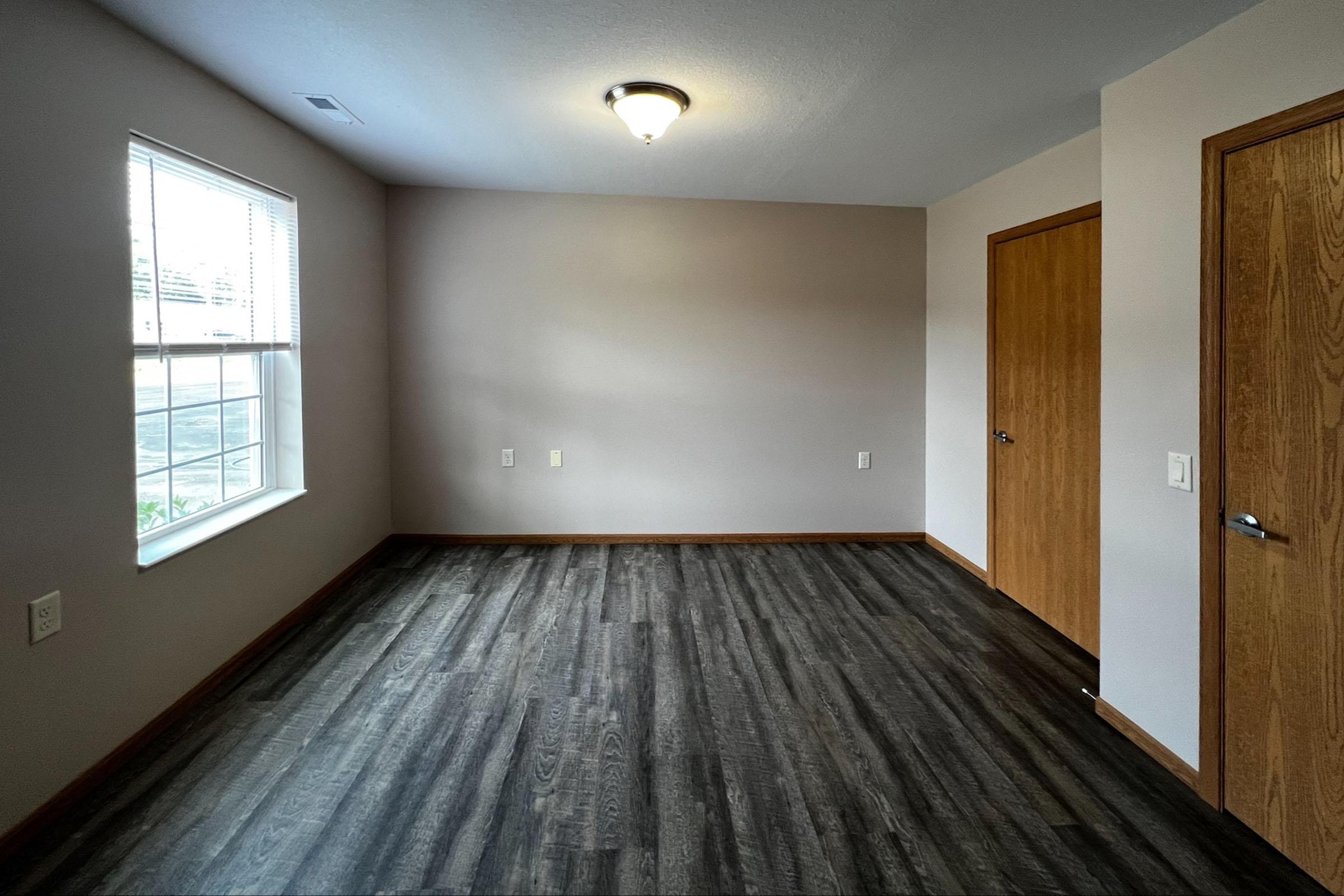 A clean, empty room featuring hardwood floors, a single window with blinds, a light fixture on the ceiling, and two doors on opposite walls. The walls are painted a light color, creating a bright and spacious atmosphere.