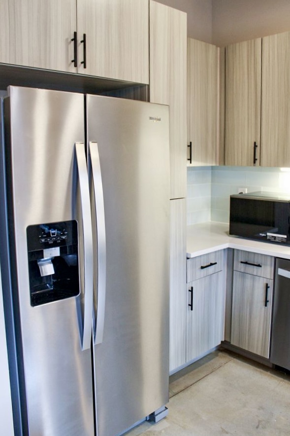 A modern kitchen featuring a stainless steel double-door refrigerator, wood-patterned cabinetry, and a countertop with a microwave. The design is sleek and contemporary, with a clean and organized appearance.