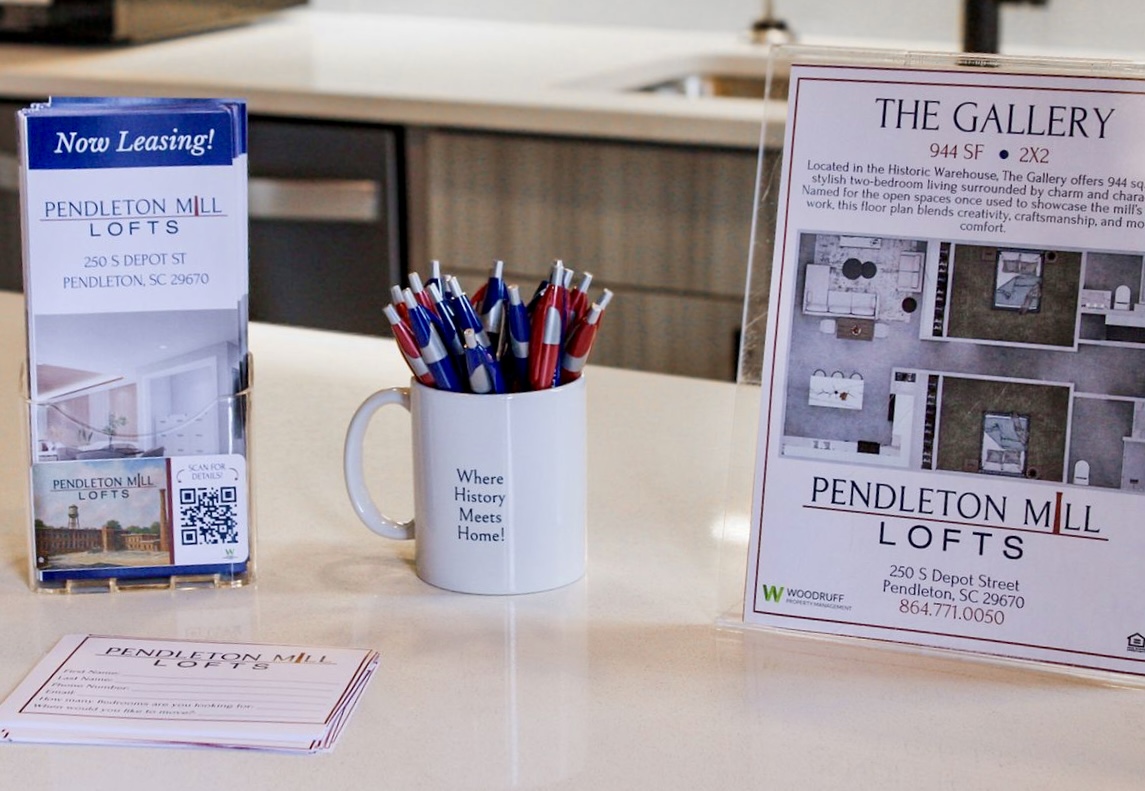 A countertop display featuring a leasing brochure for Pendleton Mill Lofts. It includes a cup filled with pens, a sign reading "Where History Meets Home," and details about available lofts, including square footage and contact information. A bright and welcoming leasing space.