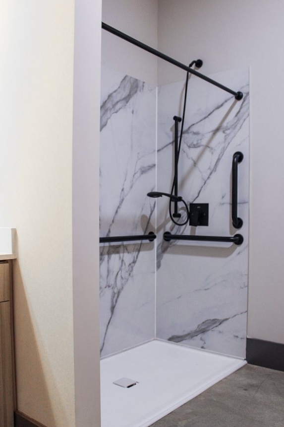 A modern bathroom shower featuring a marble-patterned wall, black grab bars, and a sleek showerhead. The shower has a minimalist design with a white floor and is designed for accessibility, incorporating safety features for ease of use.