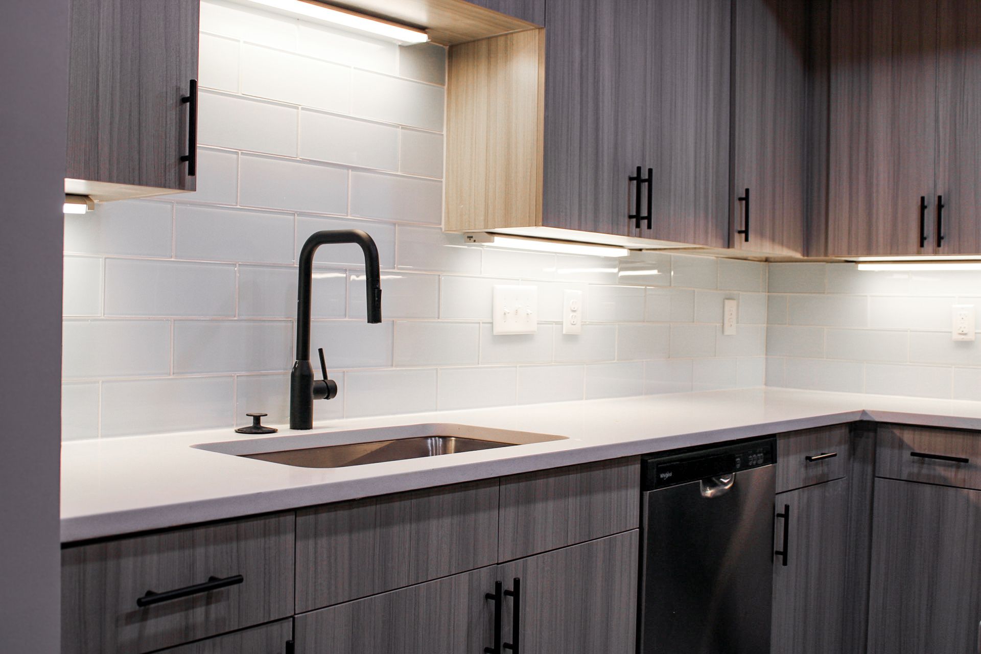 Modern kitchen countertop with a stainless steel sink, sleek black faucet, and under-cabinet lighting. The walls feature light blue subway tiles, and the cabinetry is a mix of dark and light wood finishes. A dishwasher is integrated into the cabinetry.