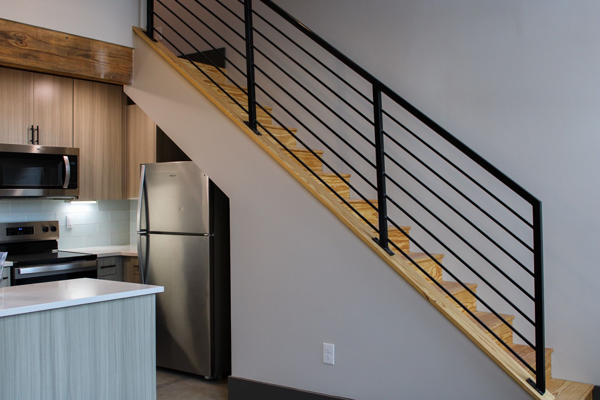 Modern interior featuring a staircase with wooden steps and black metal railing. The space includes a contemporary kitchen area with stainless steel appliances, light wood cabinetry, and a minimalist design. Natural light highlights the open layout and clean lines.