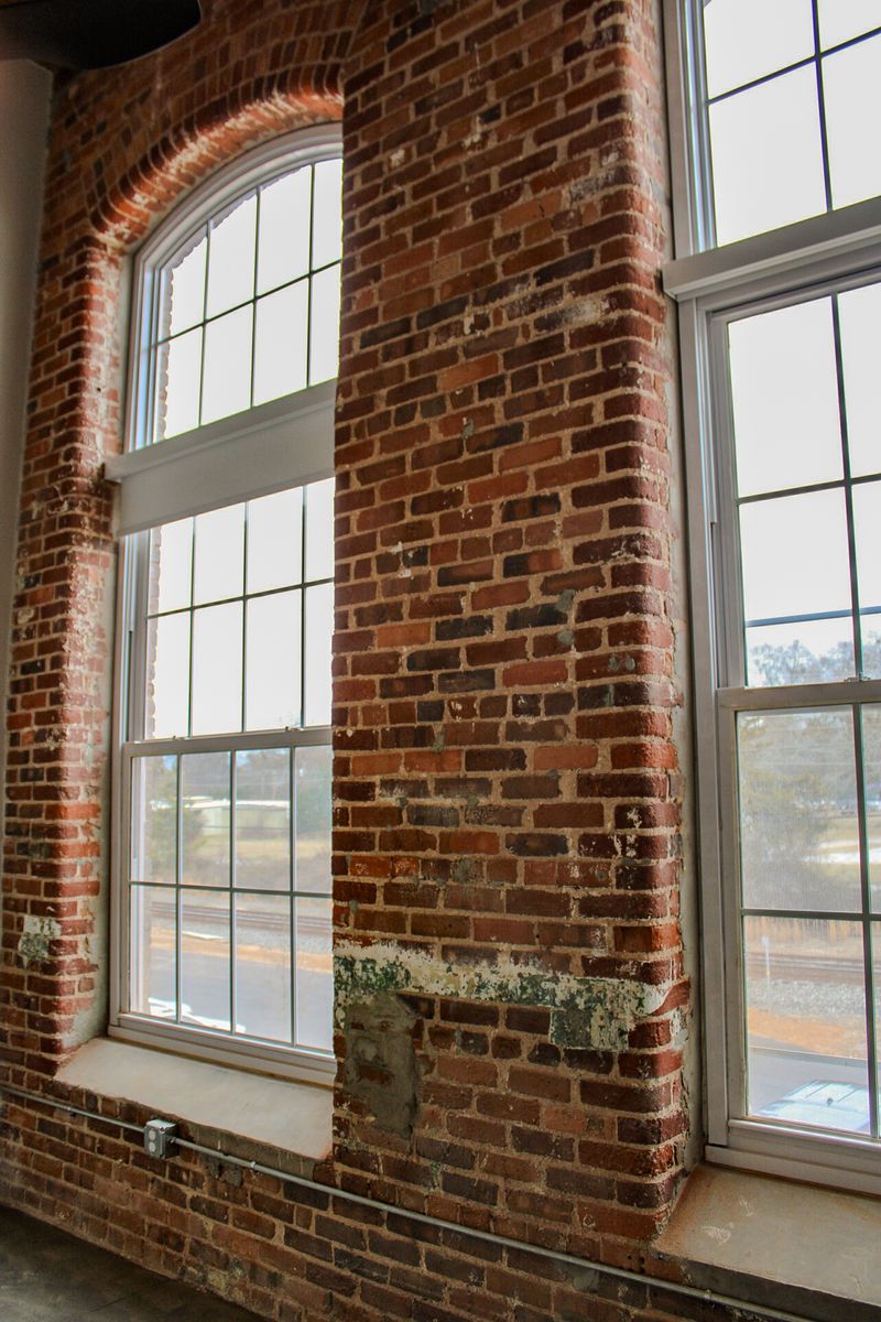 Interior view of a room featuring two large arched windows framed by exposed red brick walls. The brick has a weathered appearance, with some patches of paint and damage. Natural light filters through the windows, casting shadows on the floor.