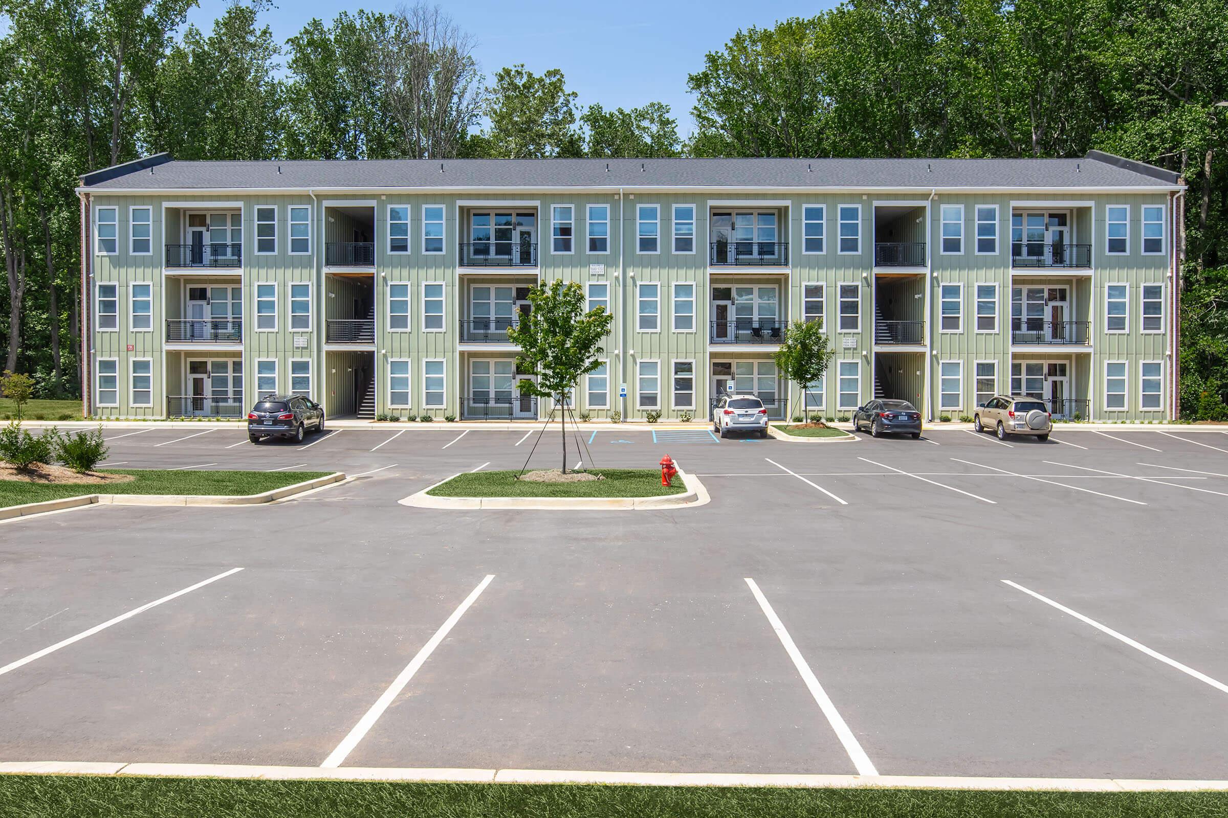 Three-story apartment building with a light green exterior, featuring balconies on each floor. The surrounding area has a well-maintained parking lot with several parked cars and green trees in the background under a clear blue sky.