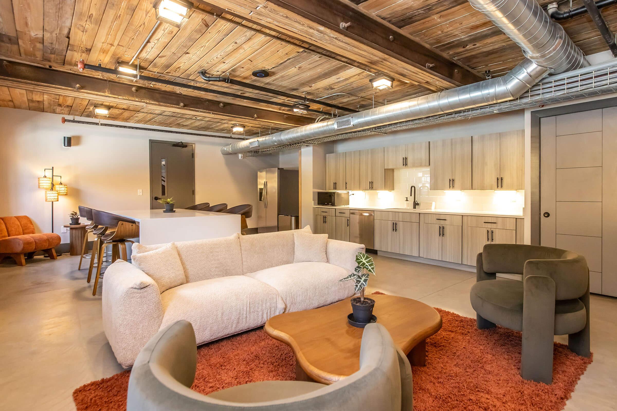 A modern living space featuring a cozy beige sofa, two green armchairs, and a wooden coffee table atop a textured orange rug. The background includes a minimalist kitchen with light wood cabinetry, a countertop, and sleek appliances, all set under industrial-style wooden beams and exposed ductwork.